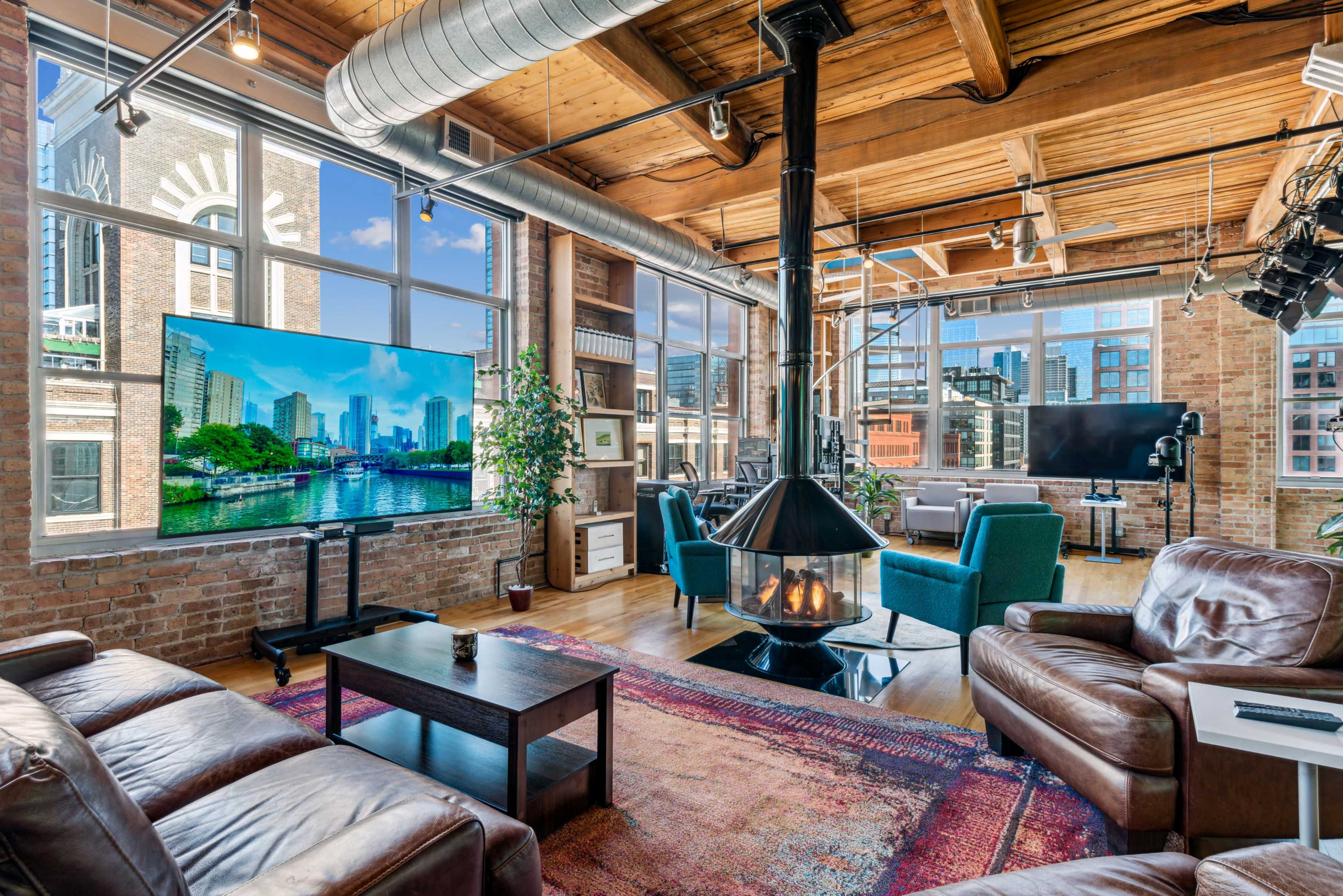 A spacious loft-style living area with large windows, a modern television, a central fireplace, and comfortable seating arrangements.