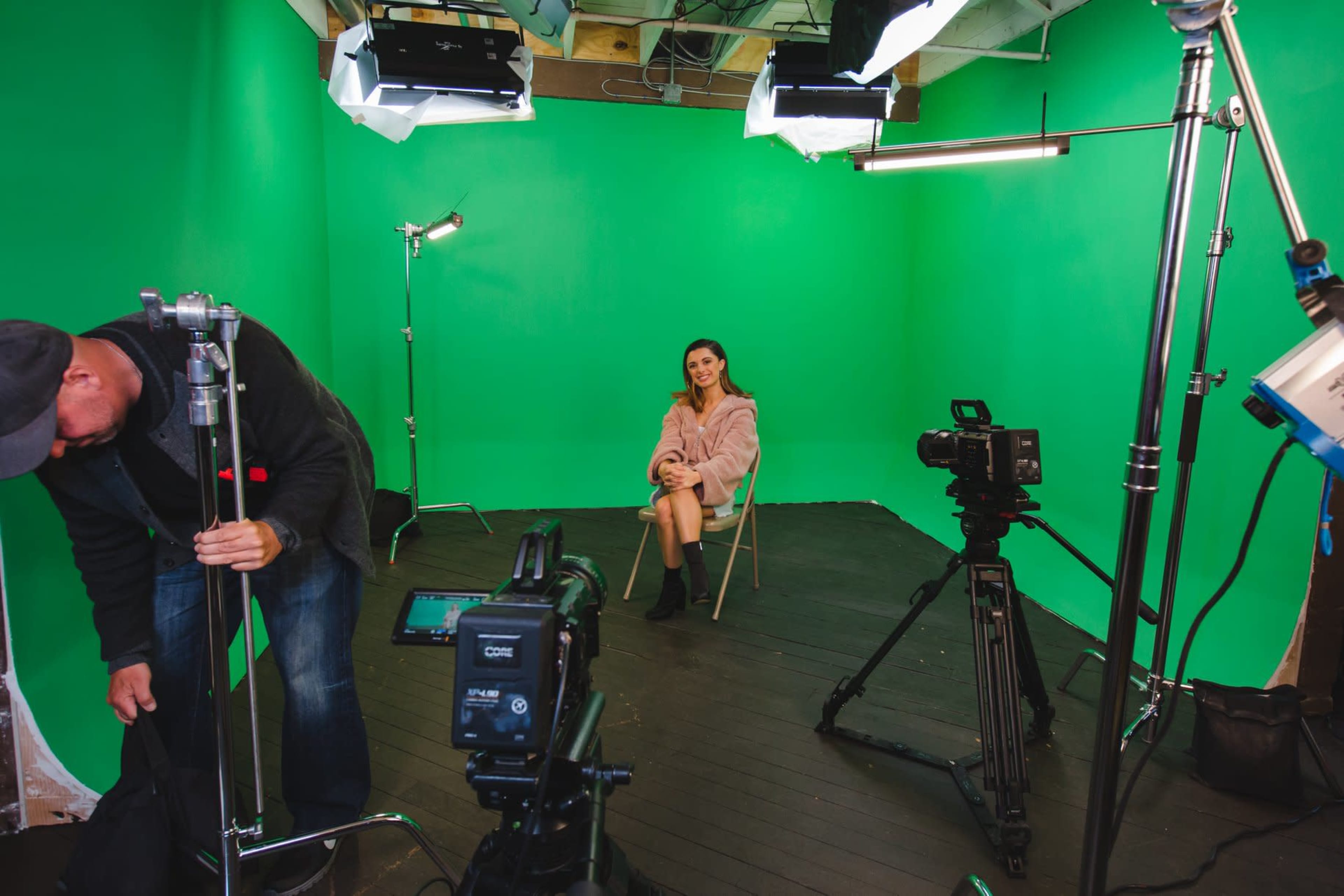 A woman sits on a chair in the center of a green screen studio, surrounded by various filming equipment and lighting.