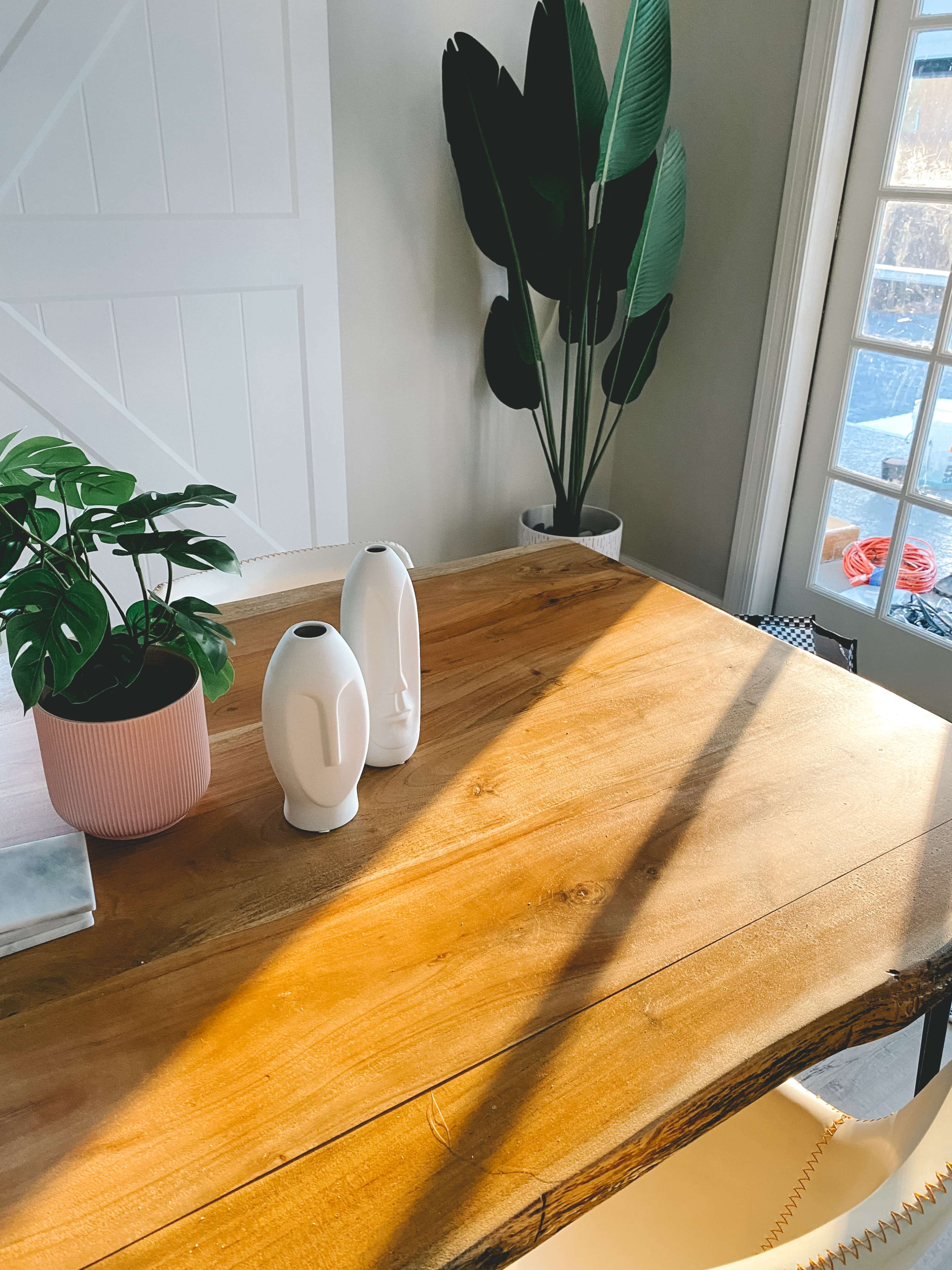 A wooden table is topped with two white vases and a potted plant, with sunlight casting shadows across the surface.