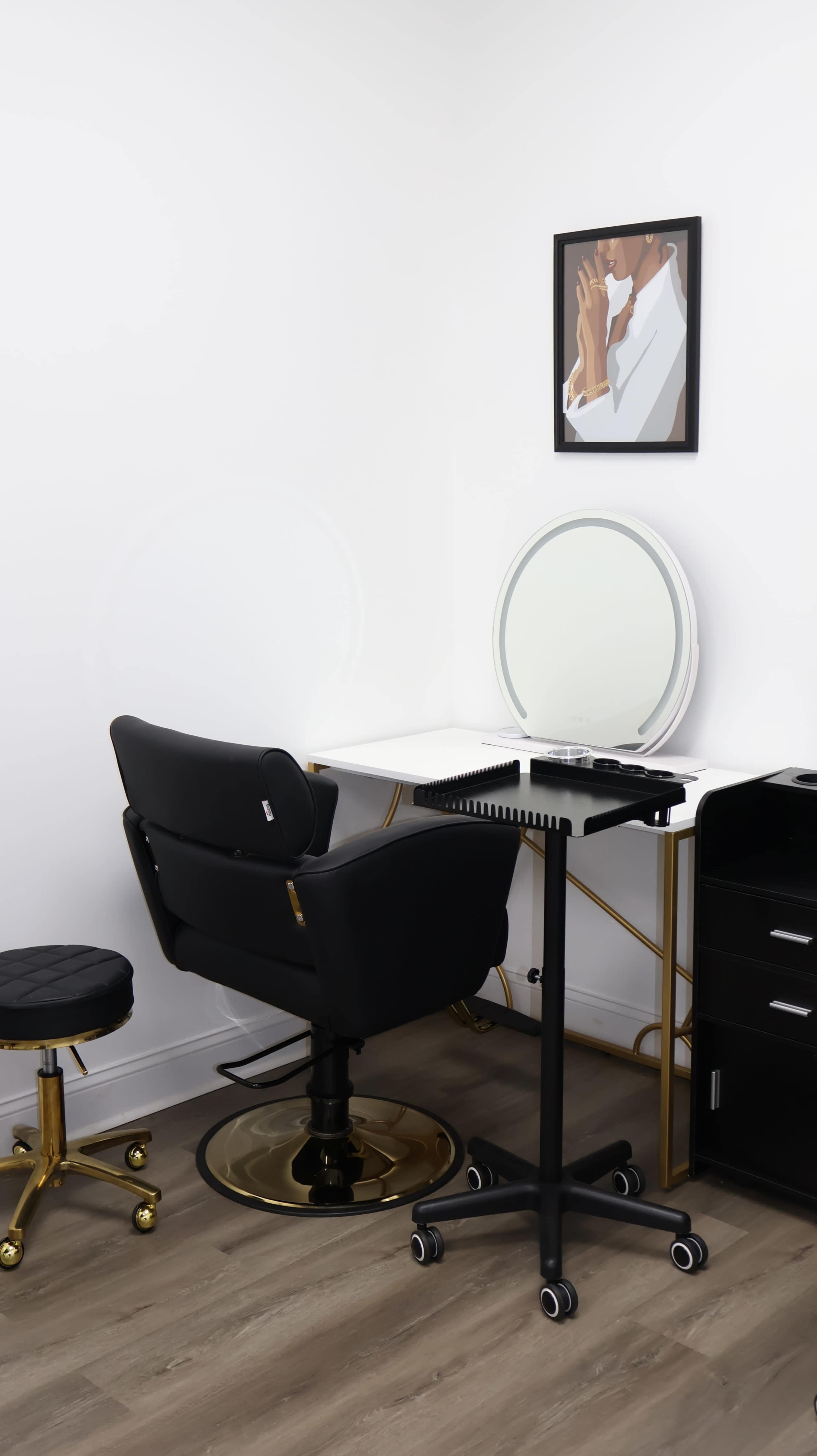 The image shows a minimalistic beauty salon space featuring a black chair, a round mirror on a white desk, a rolling cart, and a small black stool.