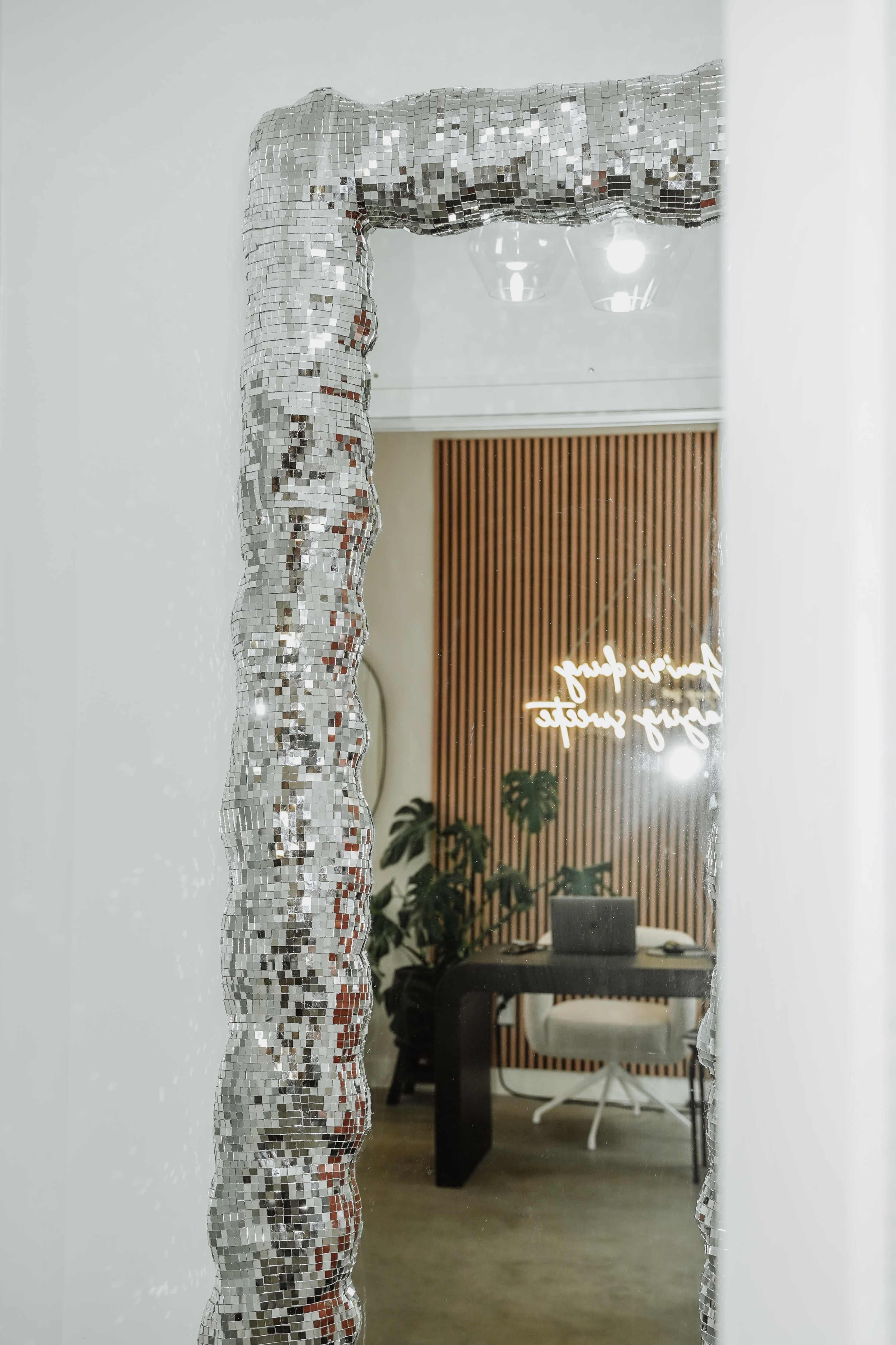 A decorative, mirrored frame surrounds a large mirror, reflecting a modern interior with wooden paneling and indoor plants.