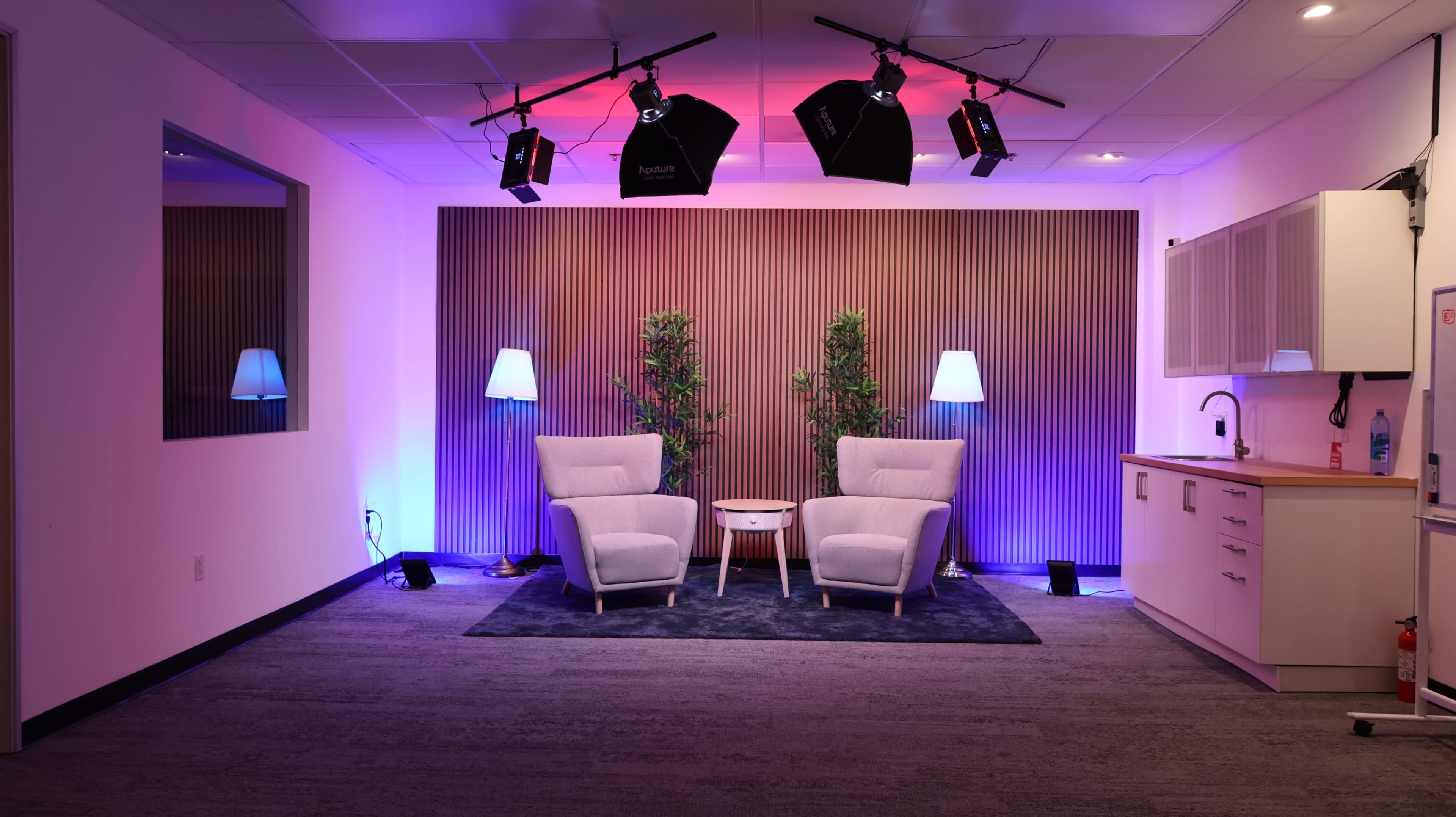 A modern studio setup with two white armchairs facing a small table, surrounded by illuminated plants and soft lighting in purple and blue tones.