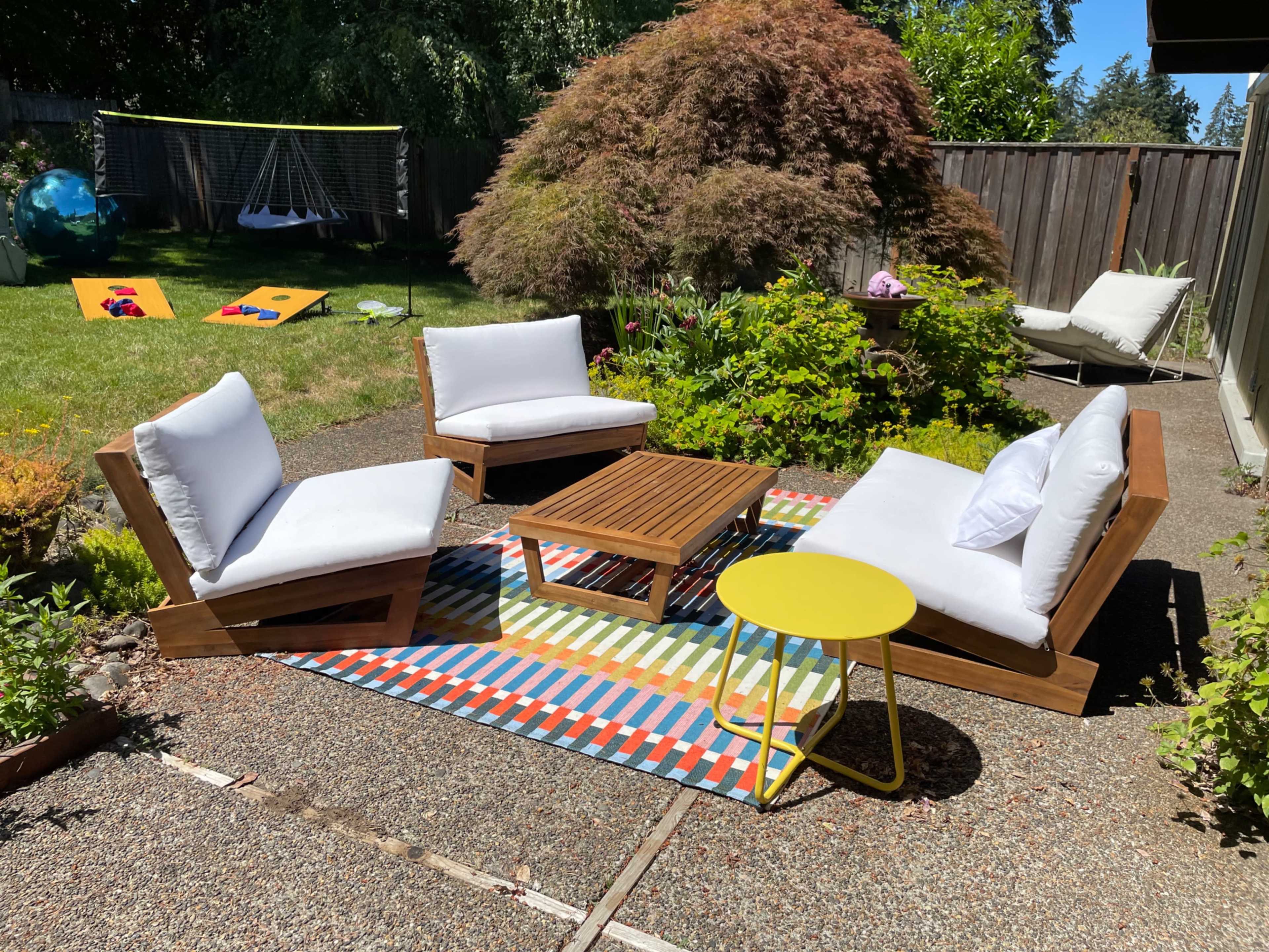 The image shows an outdoor seating area with two modern lounge chairs and a small table on a colorful patterned rug, surrounded by greenery and a wooden fence.