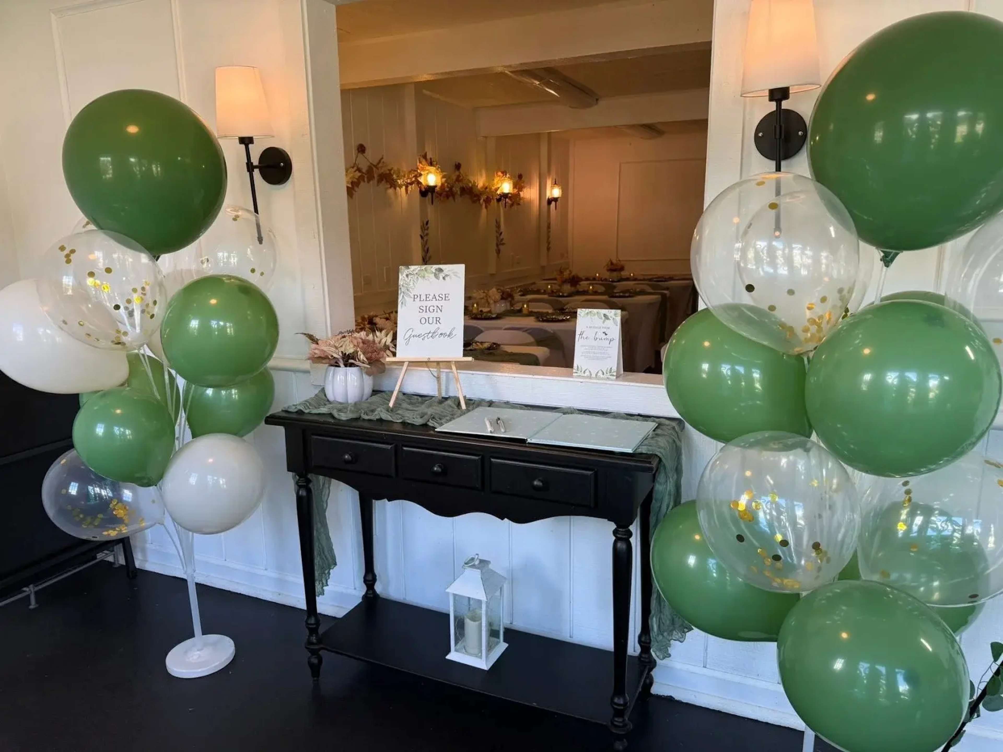 The image shows a reception area decorated with green and white balloons, a black table with a guest sign-in sheet, and a sign that reads "Please sign our guestbook."