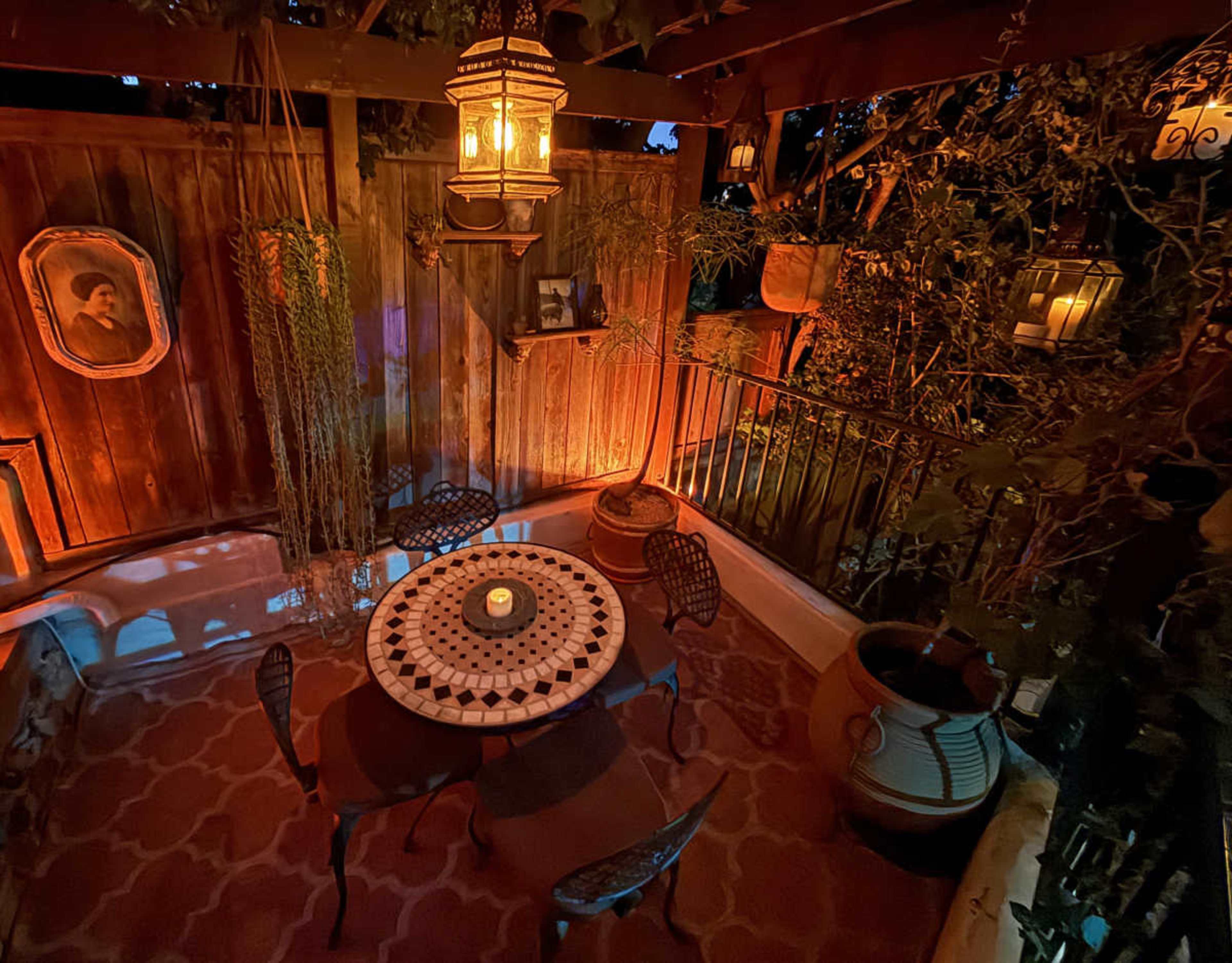 The image shows a cozy outdoor dining area with a circular table, four chairs, and decorative lanterns hanging from the wooden structure, surrounded by plants and a wall adorned with artwork.