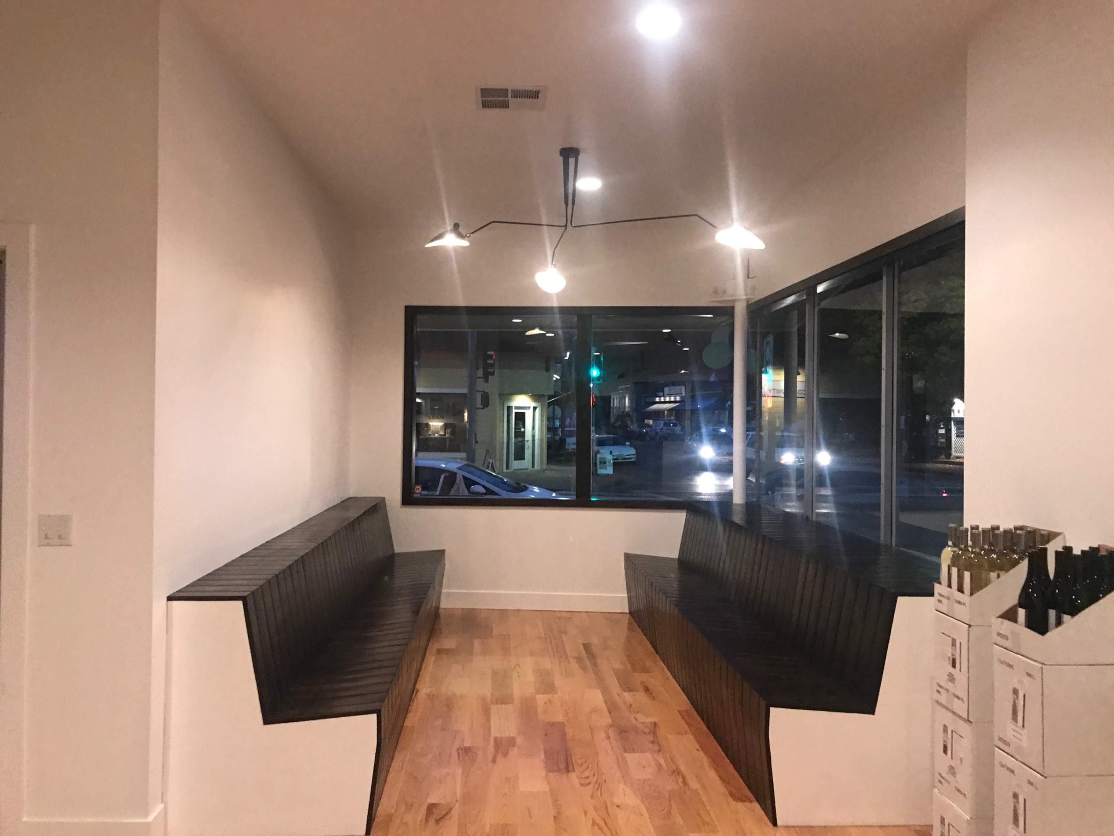 A minimalist interior features two black wooden benches facing a large window with street view and modern light fixtures overhead.
