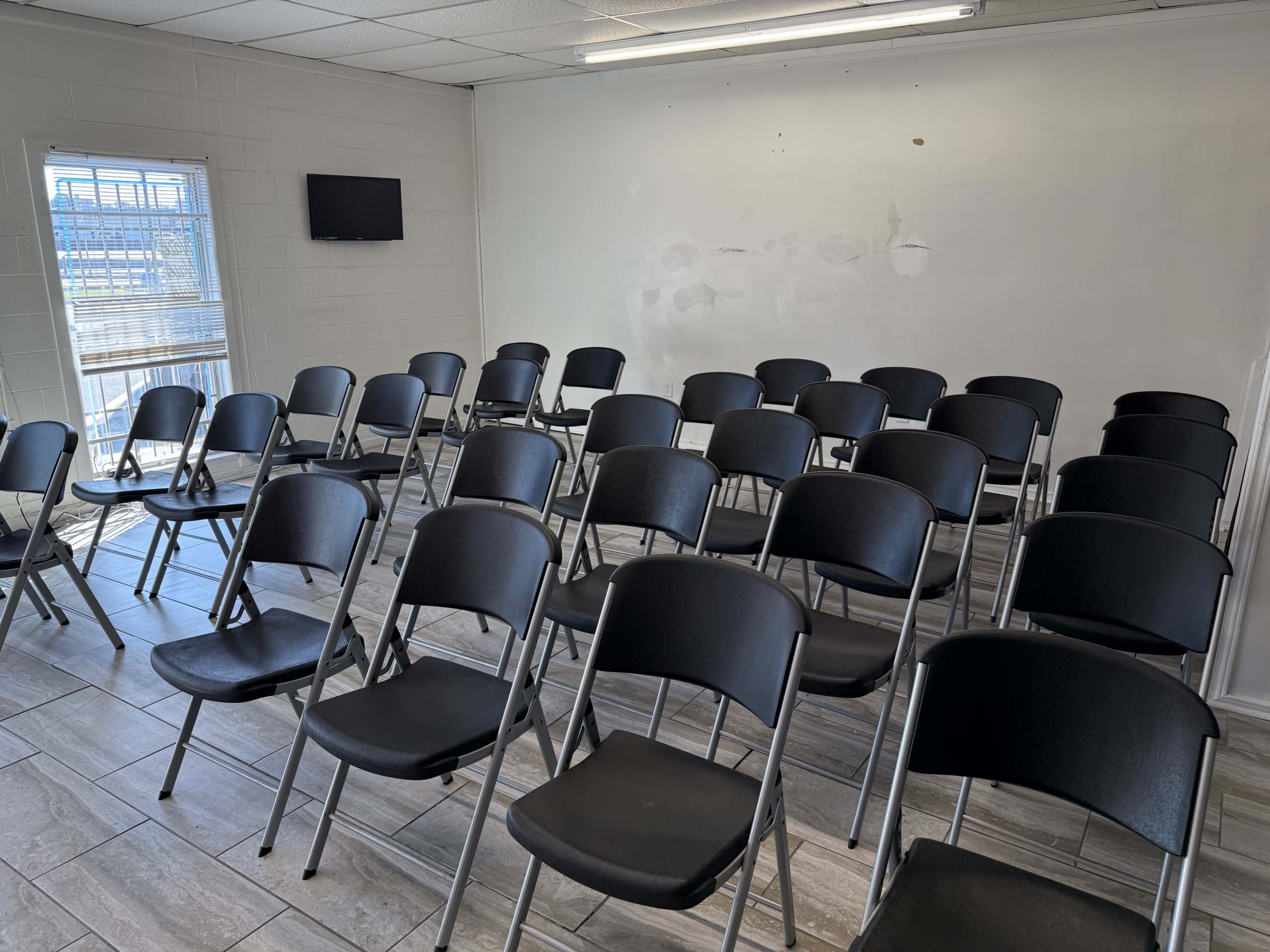 The image shows a room with rows of black folding chairs arranged facing a blank wall, with a small television mounted on the opposite wall.
