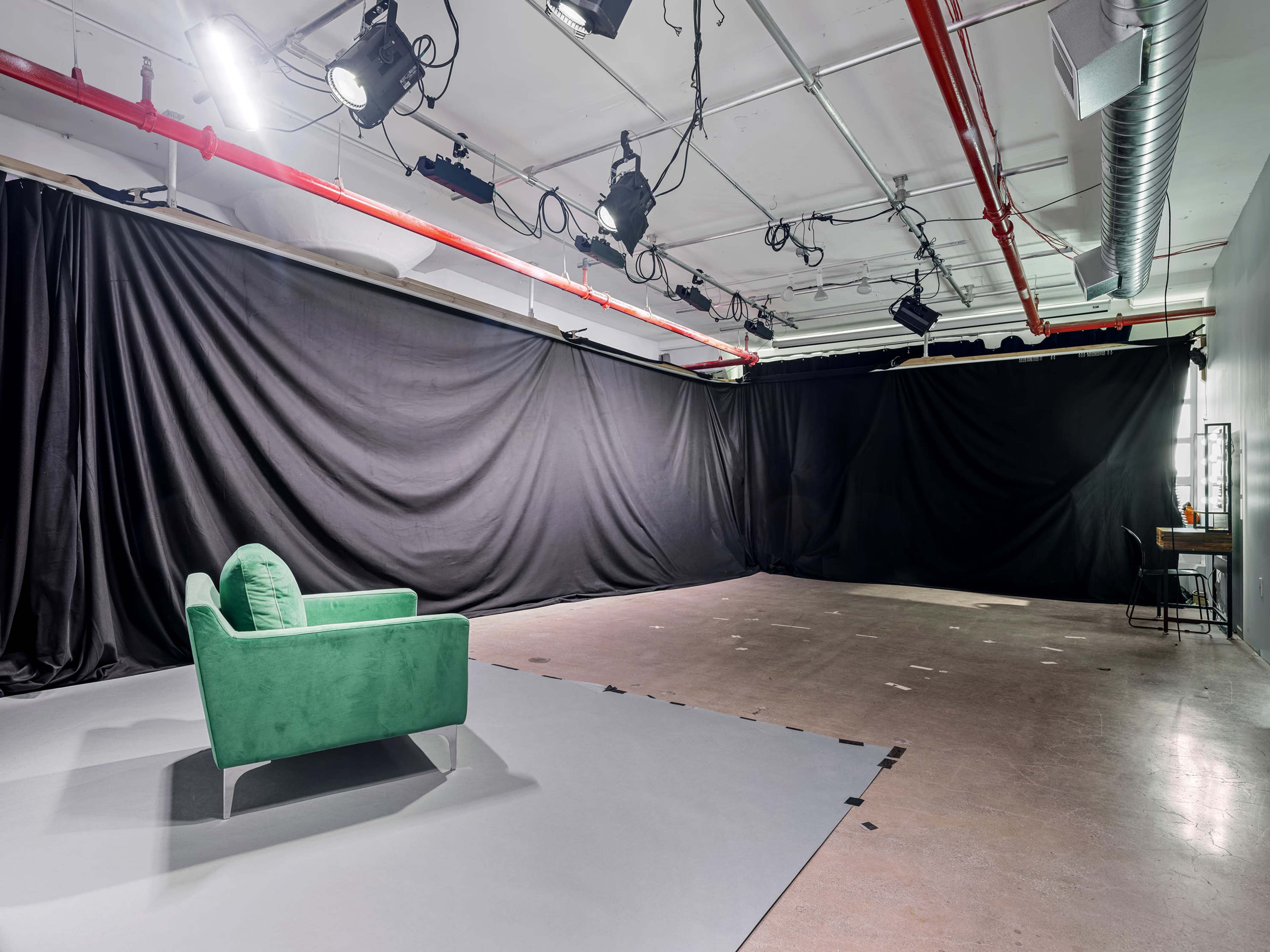 The image shows an empty studio space with a green armchair positioned on a gray floor, surrounded by black curtains and overhead lighting.