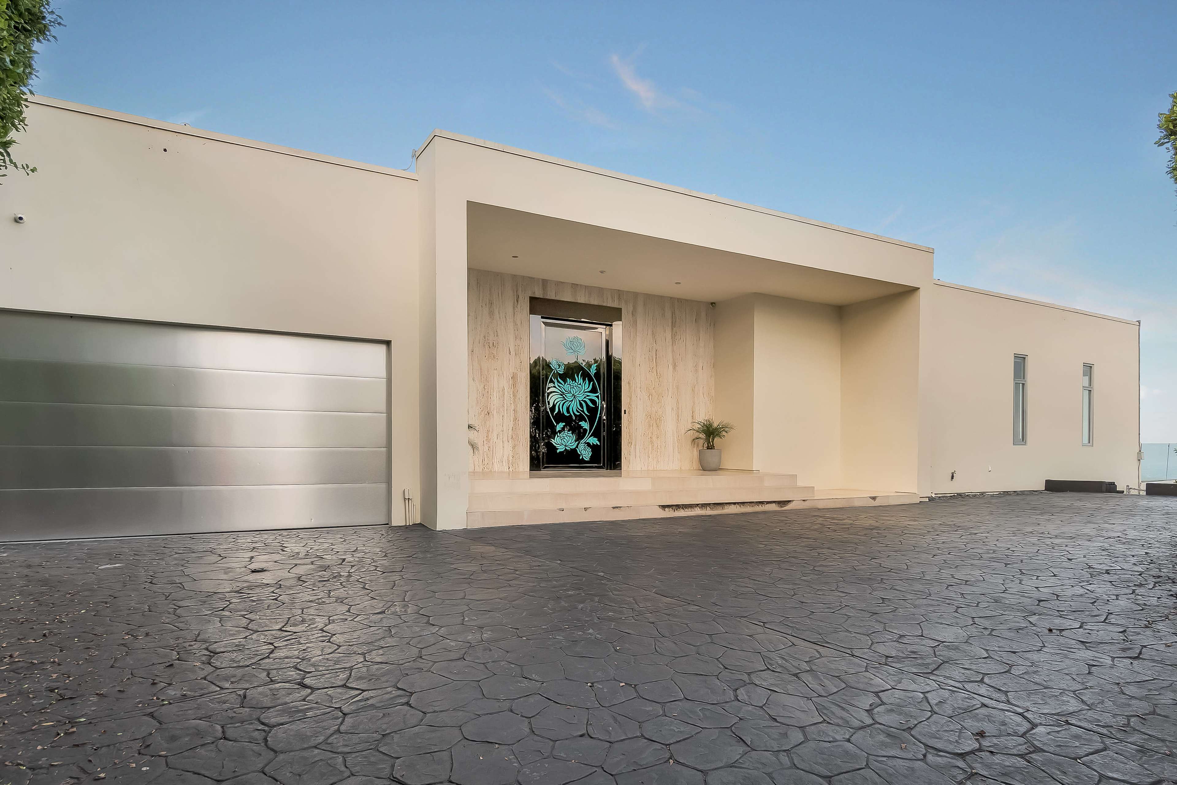 The image shows a modern, minimalist house with a large entrance and a driveway made of dark, textured material.