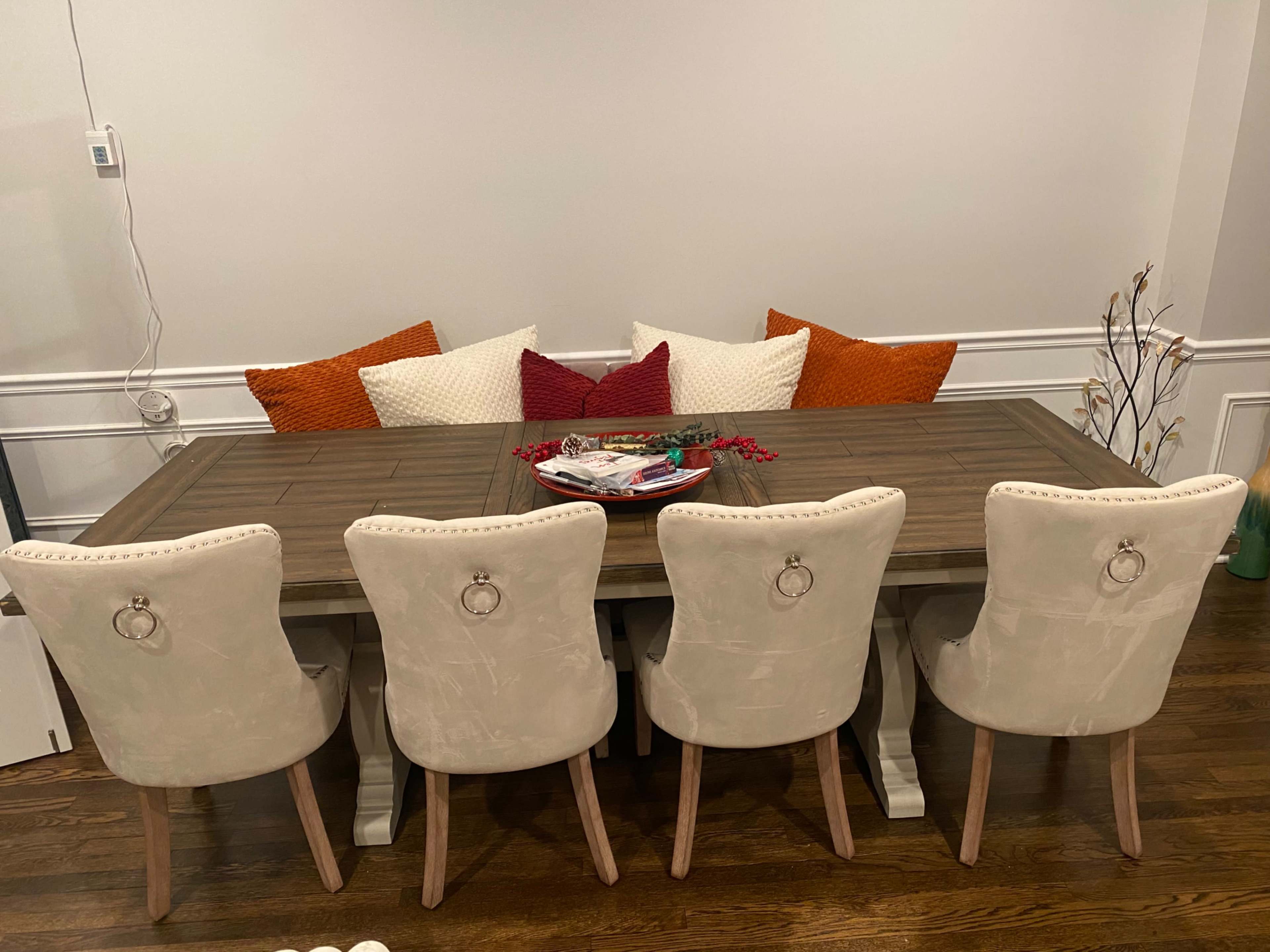 A wooden dining table with a decorative centerpiece is surrounded by four upholstered chairs, backed by colorful cushions against the wall.