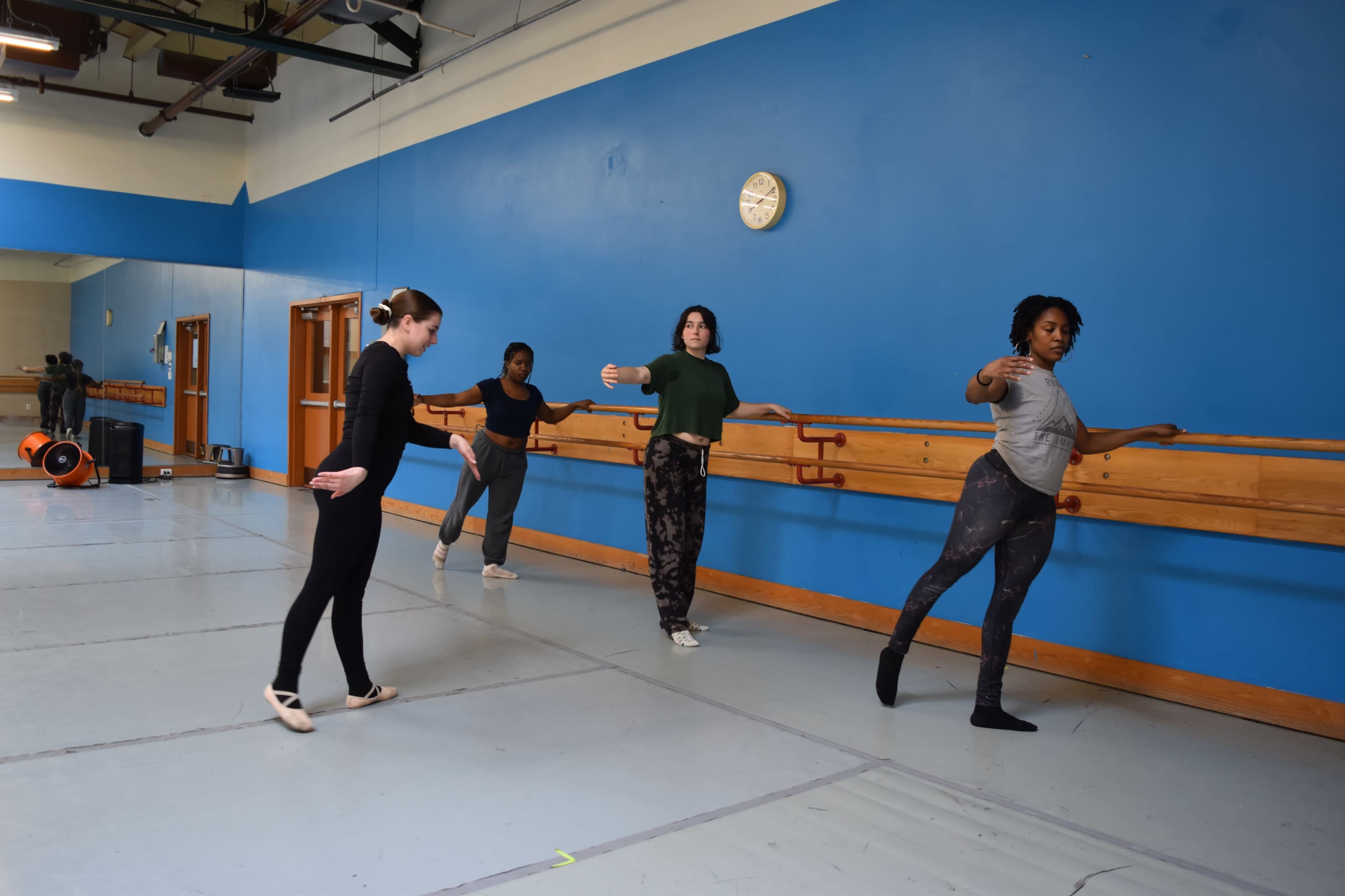 Four dancers practice ballet at a dance studio, leaning on a wooden barre with mirrors and a clock visible in the background.