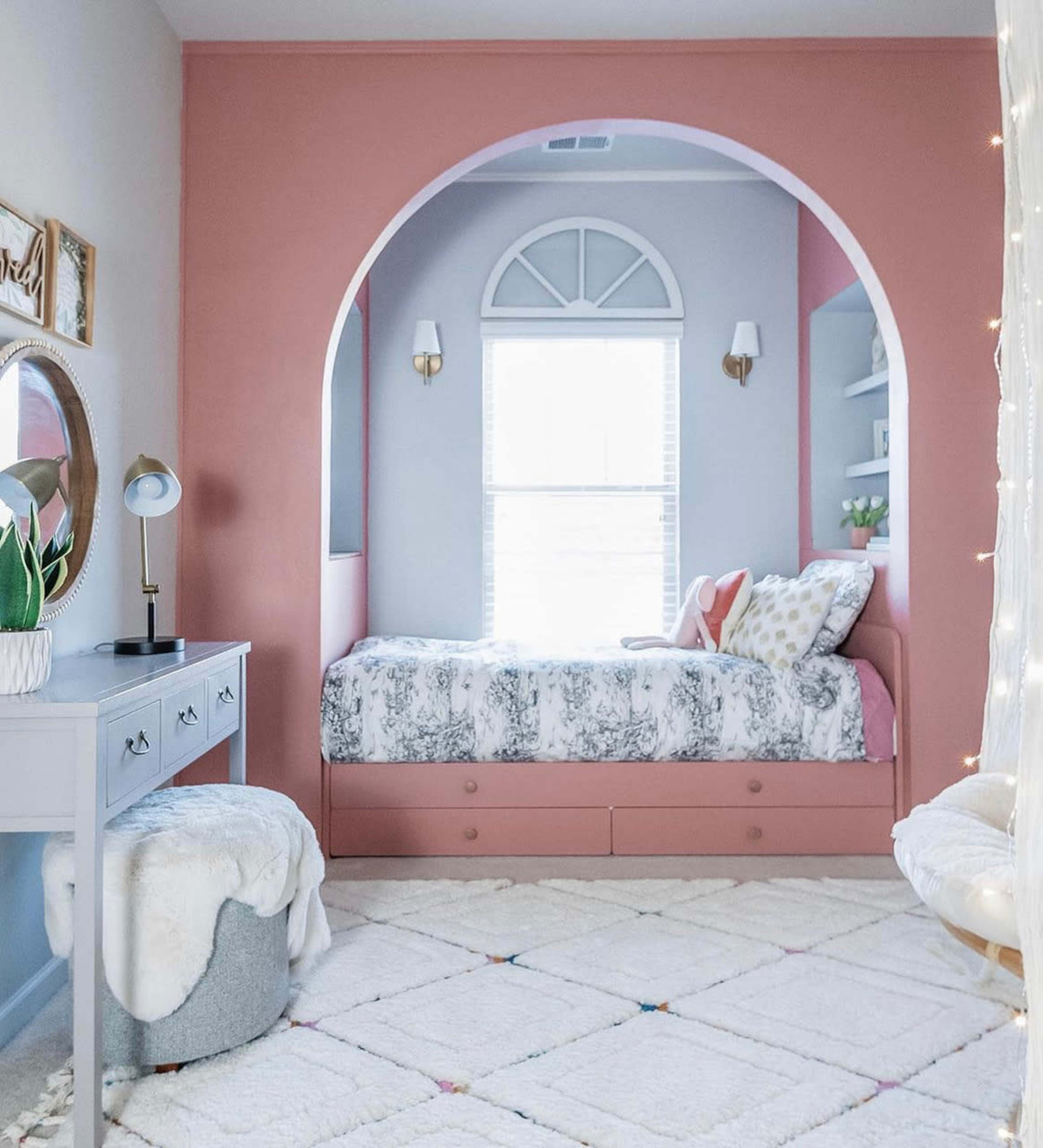 A styled bedroom features a bed in a niche with a pink archway, a gray desk, and a cozy rug.