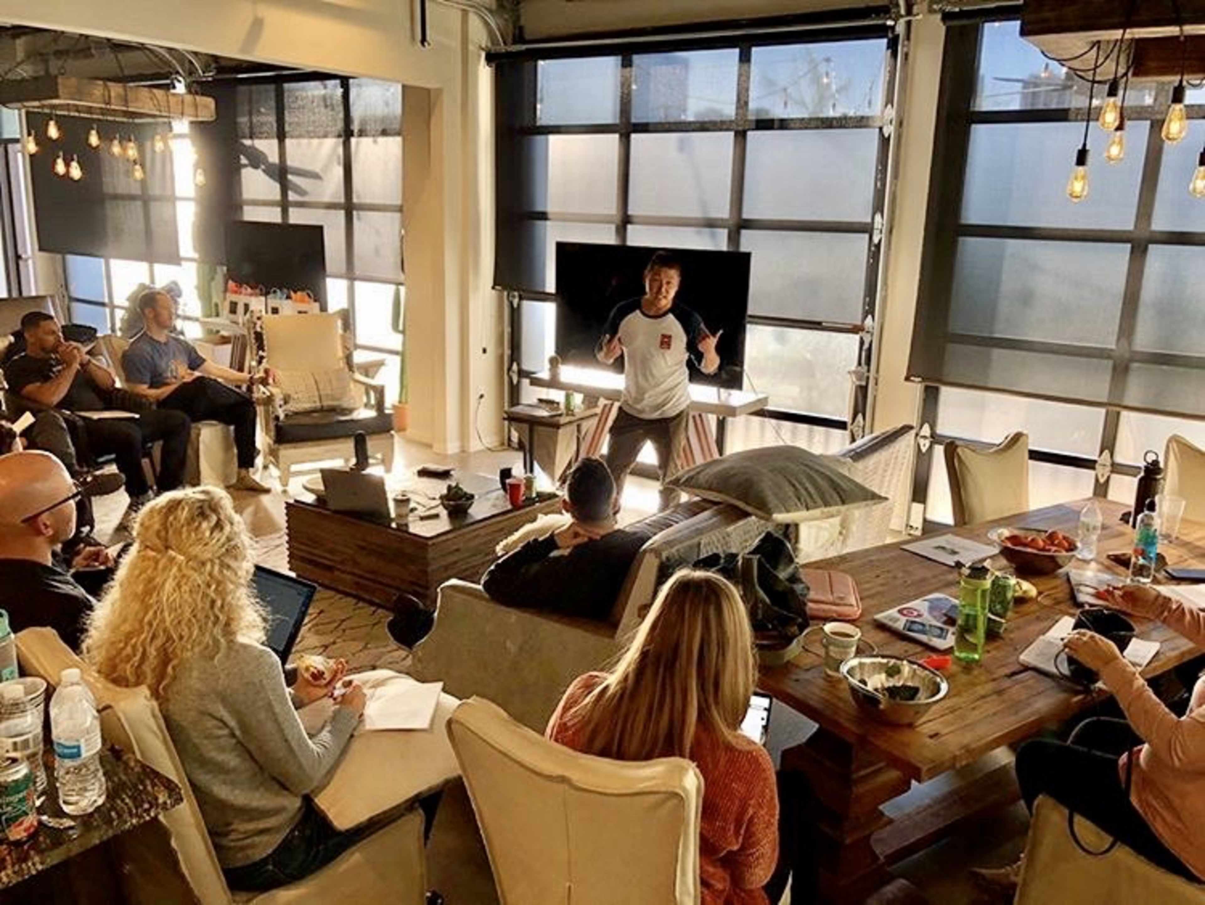 A group of people sit in a modern, well-lit space with large windows, while a speaker stands and presents to the audience.