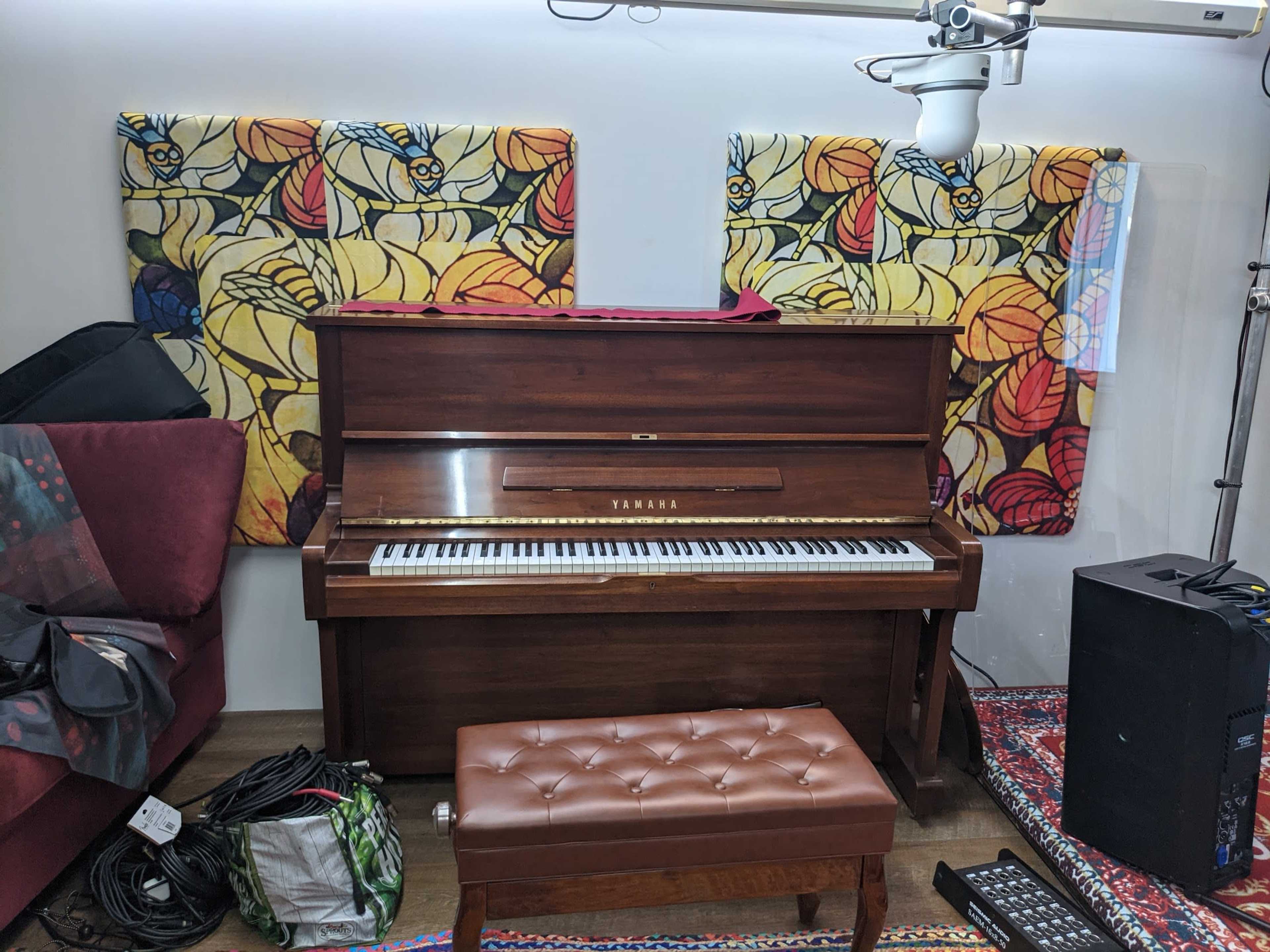 A wooden Yamaha piano situated in a room with colorful fabric wall hangings and various music equipment around it.