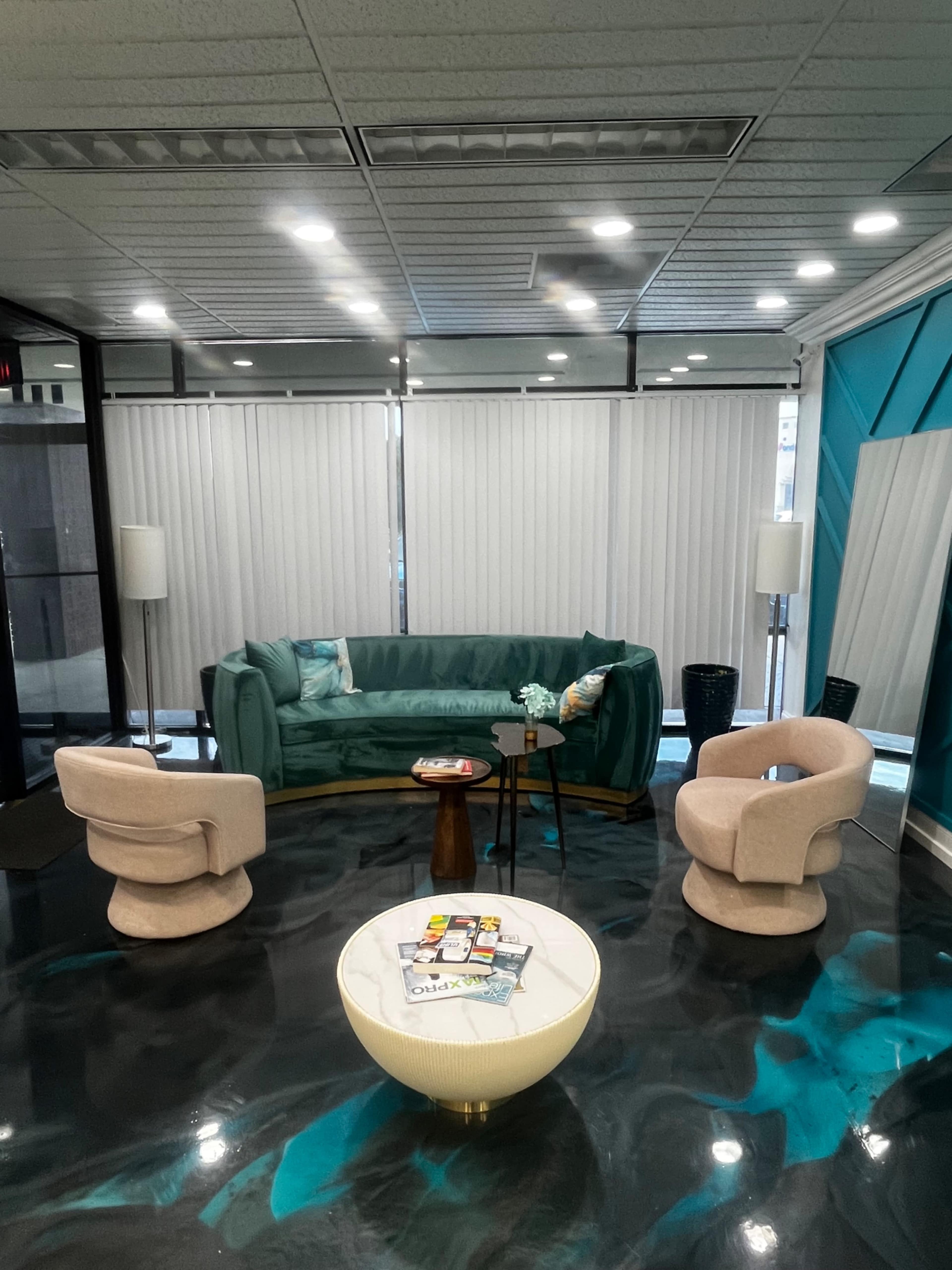 The image shows a modern waiting area with a teal couch, two light-colored armchairs, a round coffee table, and large windows covered with blinds.
