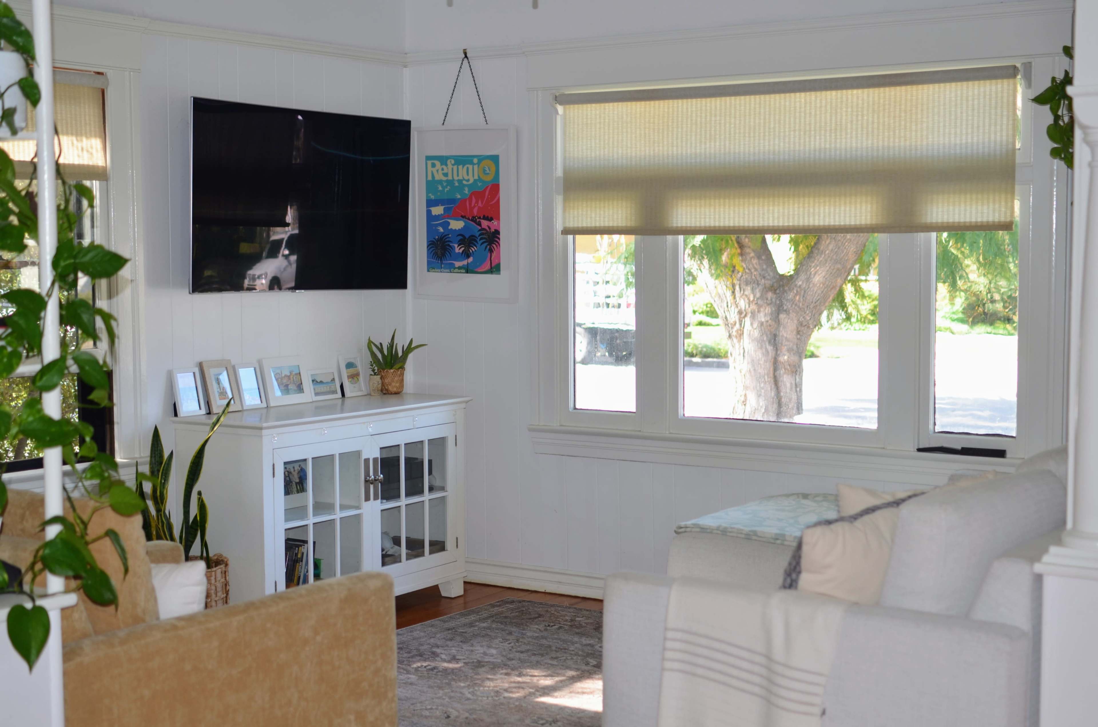 A cozy living room features a television mounted on the wall, a white cabinet with framed photos, a large window covered by a blind, and a couch with decorative pillows.