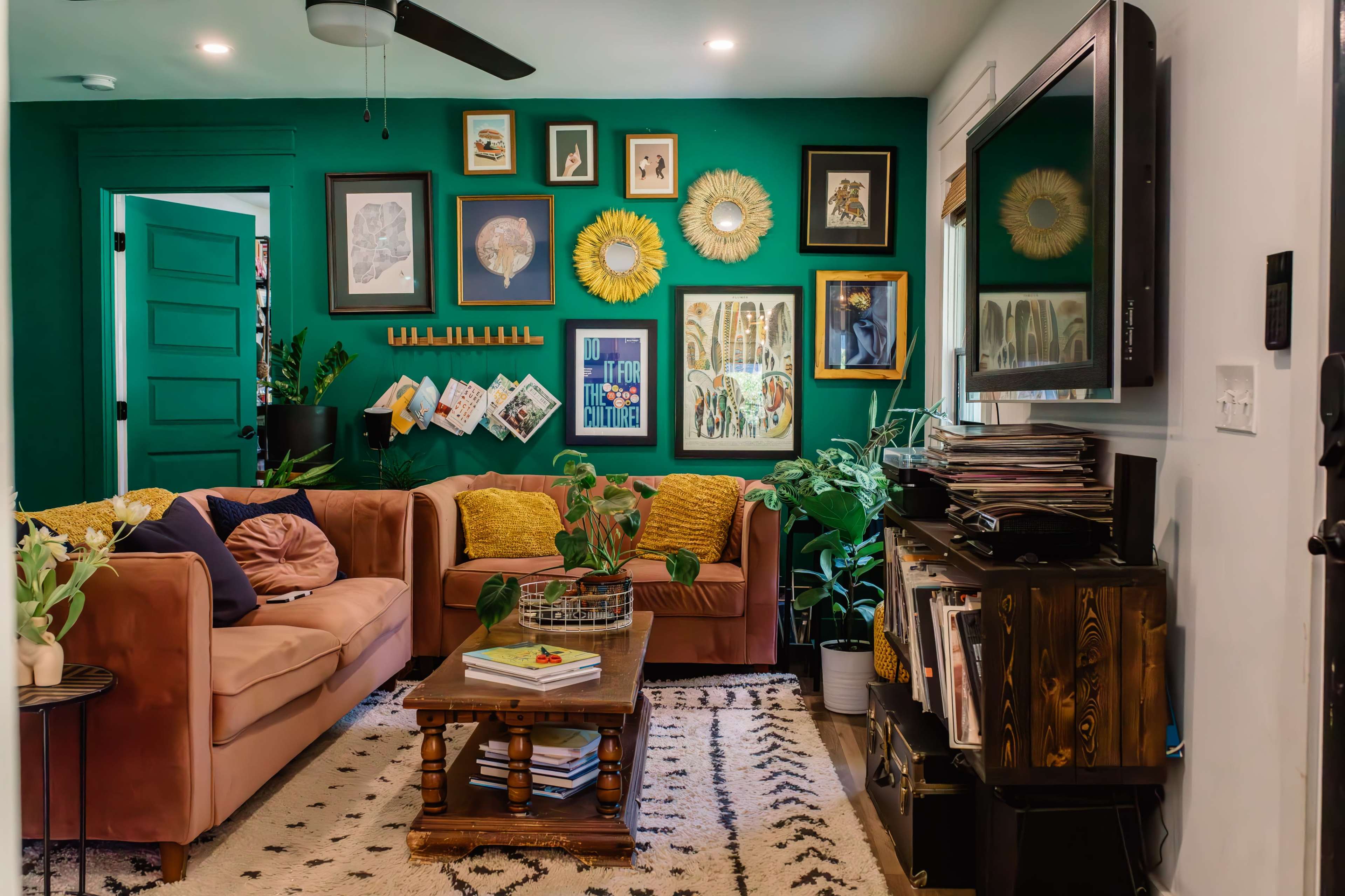 A living room features a green accent wall adorned with framed artwork, a pink sofa, a wooden coffee table, and various houseplants.