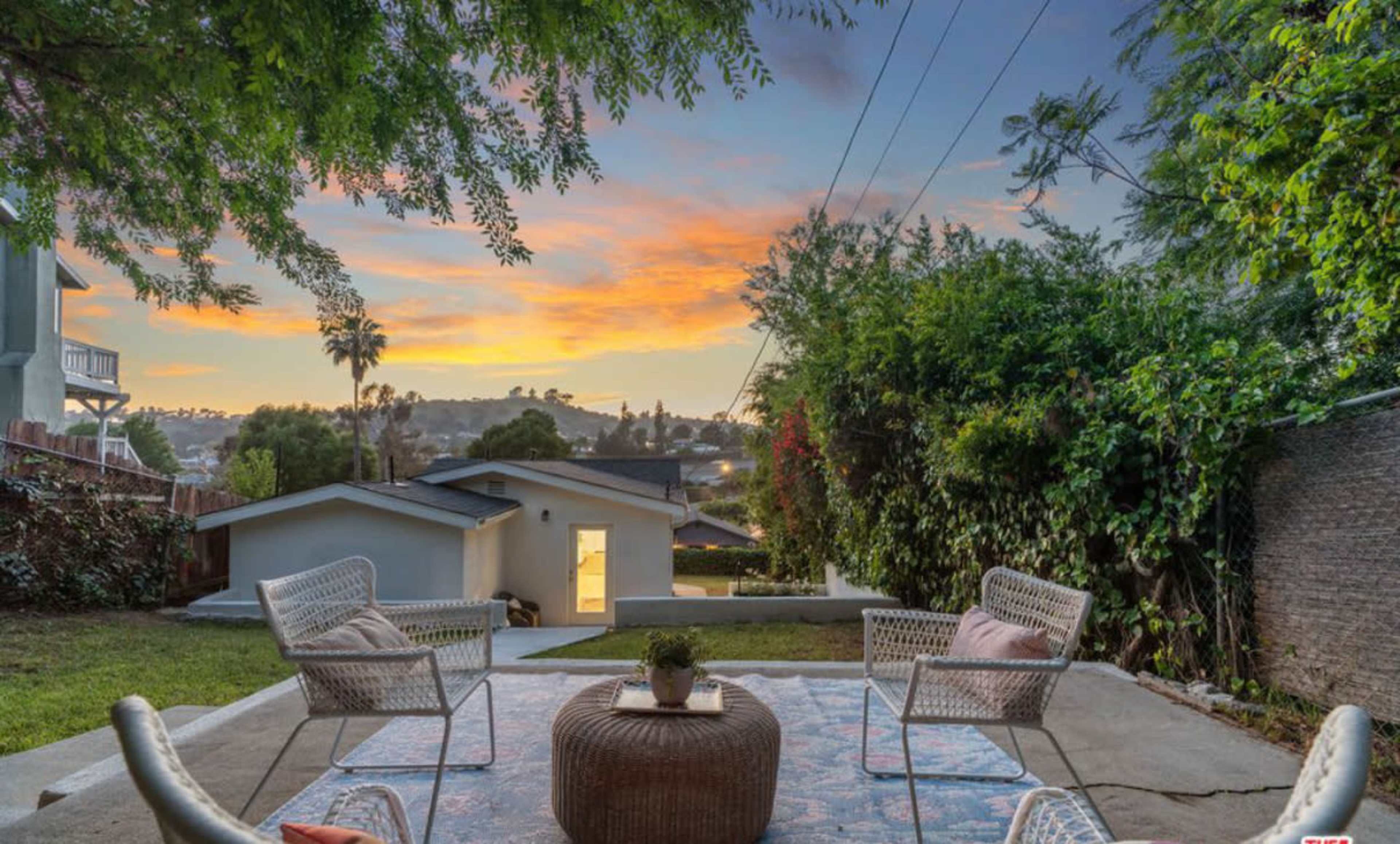 The image shows a backyard with two white chairs facing a colorful sunset over a neighborhood, framed by trees and power lines.