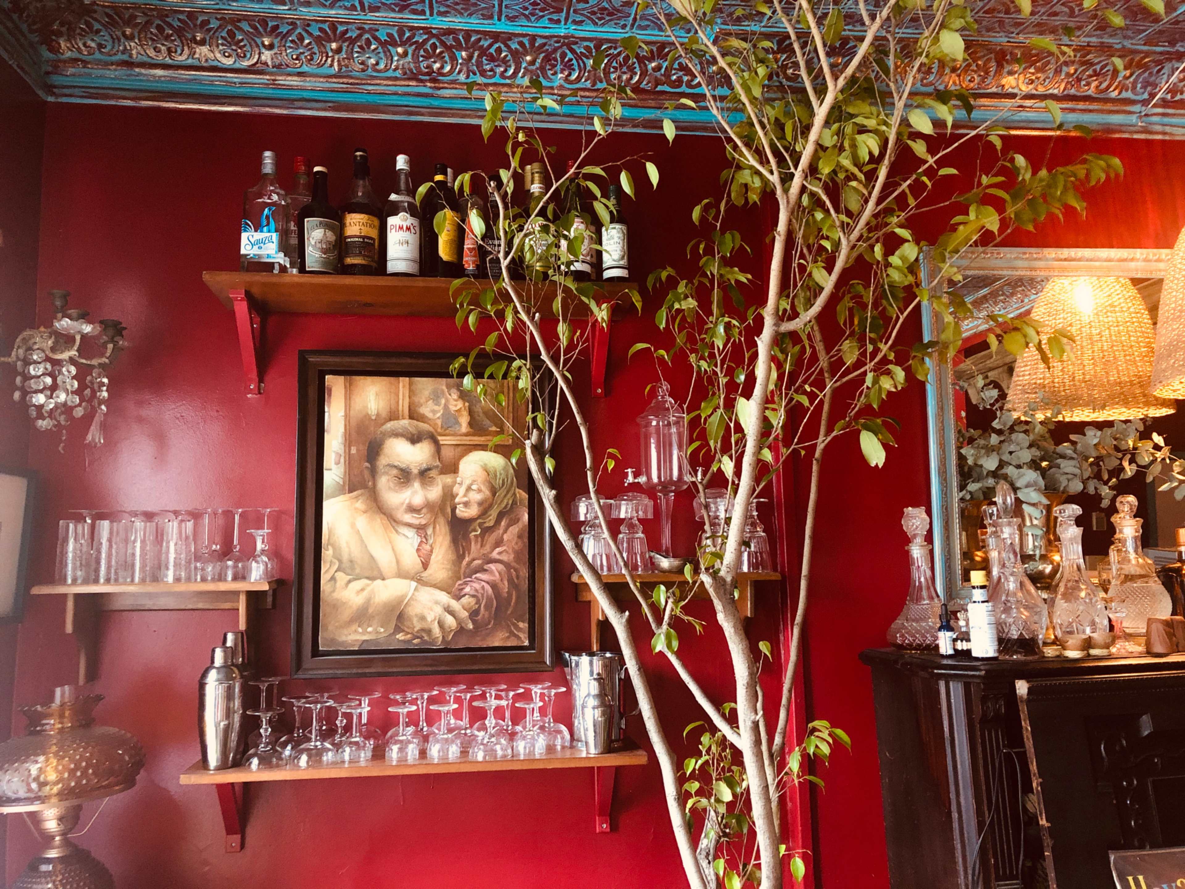 The image shows a dimly lit bar area with shelves containing bottles, glasses, and a framed painting of a man and woman on a red wall, alongside a small tree.