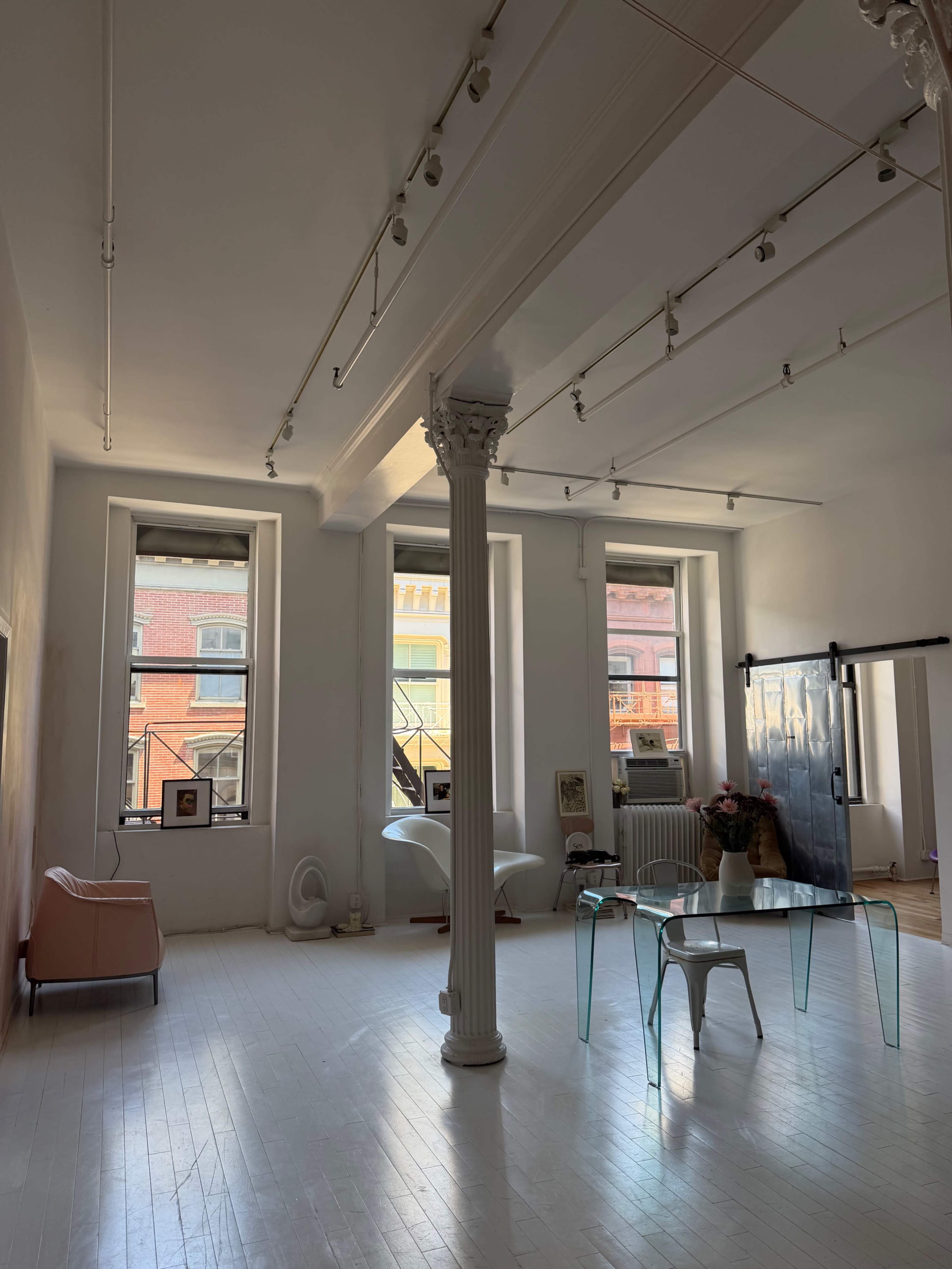 The image shows a spacious, modern room with large windows, a crystal-clear table, a minimalist couch, and a decorative column in the center.