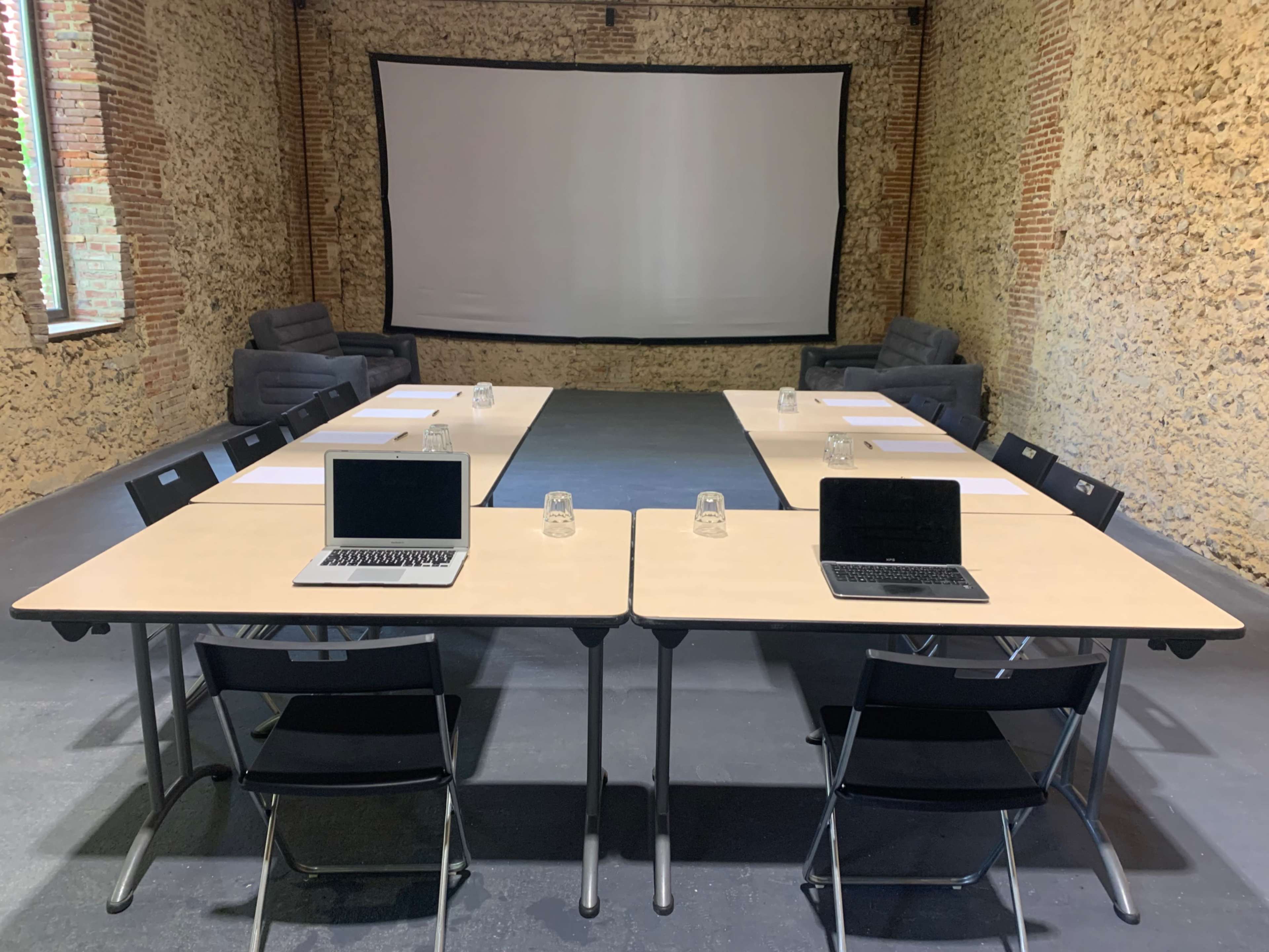 A conference room with two long tables set up in a U-shape, each equipped with laptops and glass containers, facing a large projection screen.