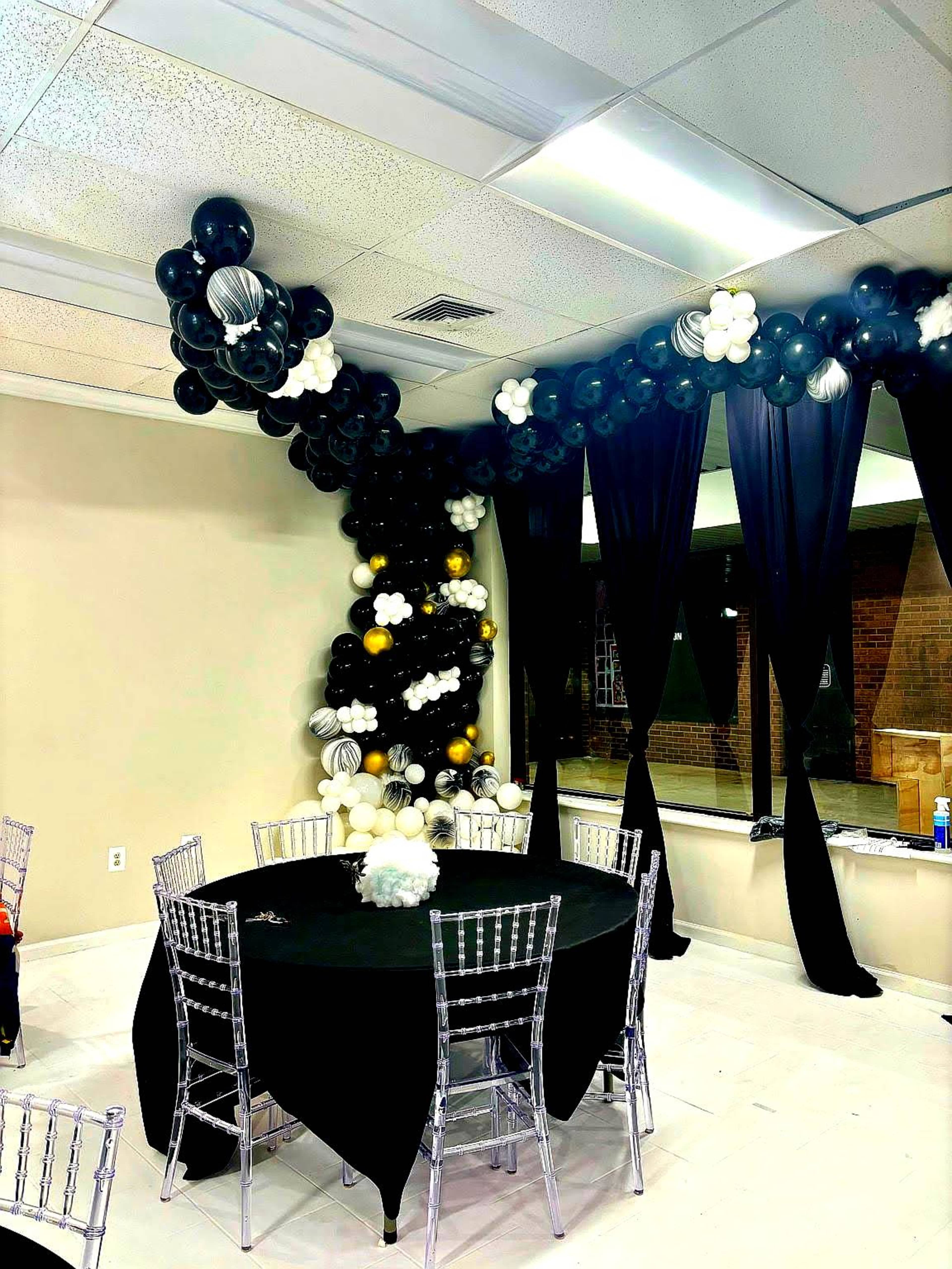 A decorated event space features a table with black cloth and silver chairs, surrounded by a balloon arch of black, white, and gold balloons.