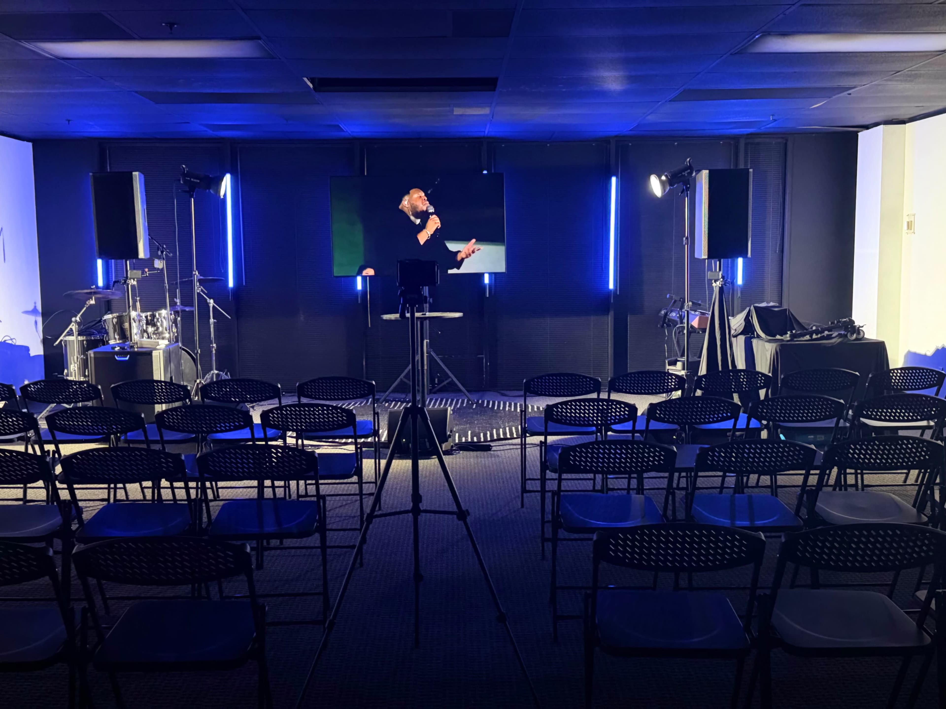 A speaker stands on a stage in a dimly lit room filled with rows of empty chairs facing a backdrop with lights and audio equipment.