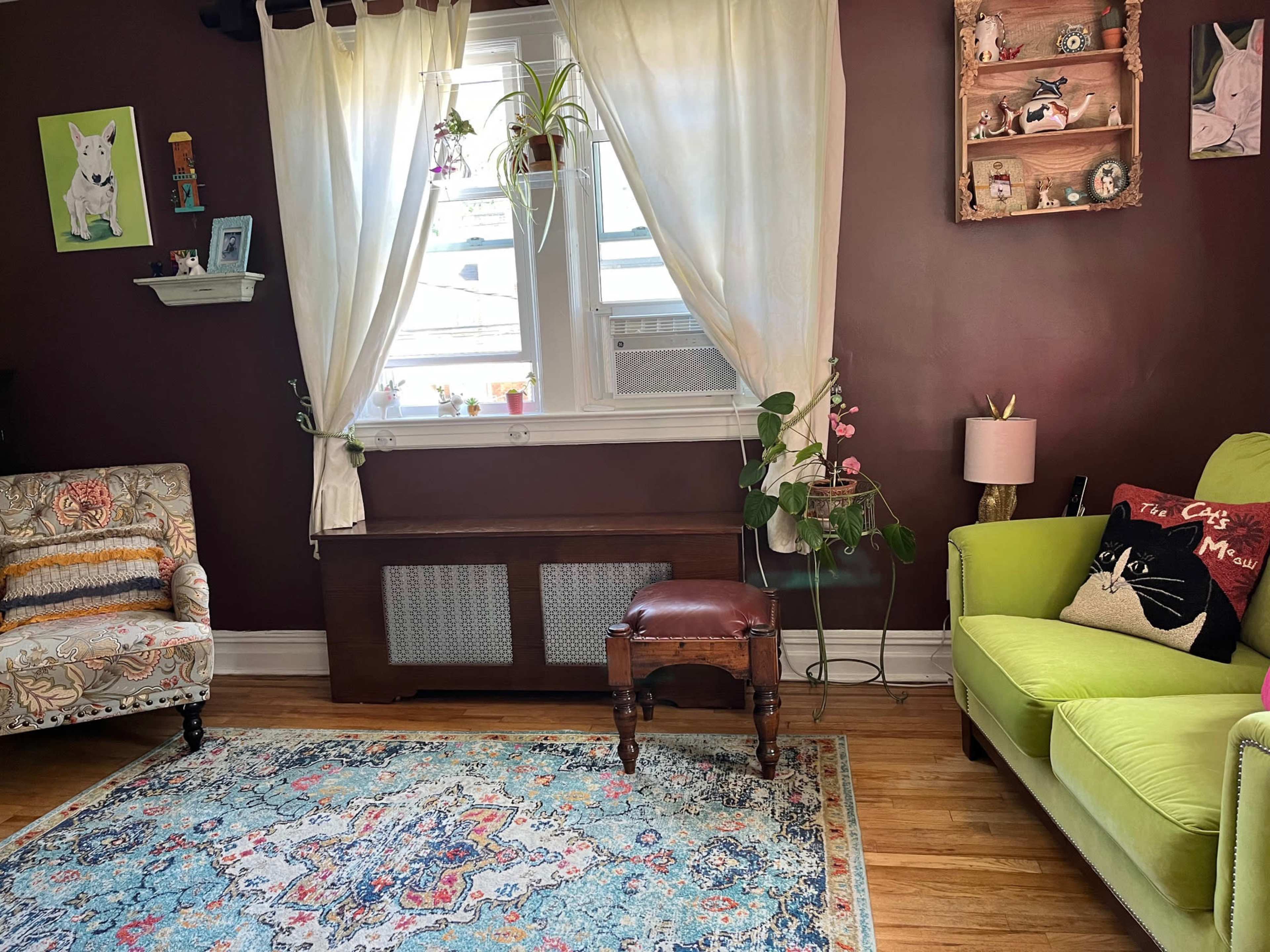 The image shows a cozy living room featuring a green couch, a patterned armchair, a window with white curtains, and decorative artwork on the walls.