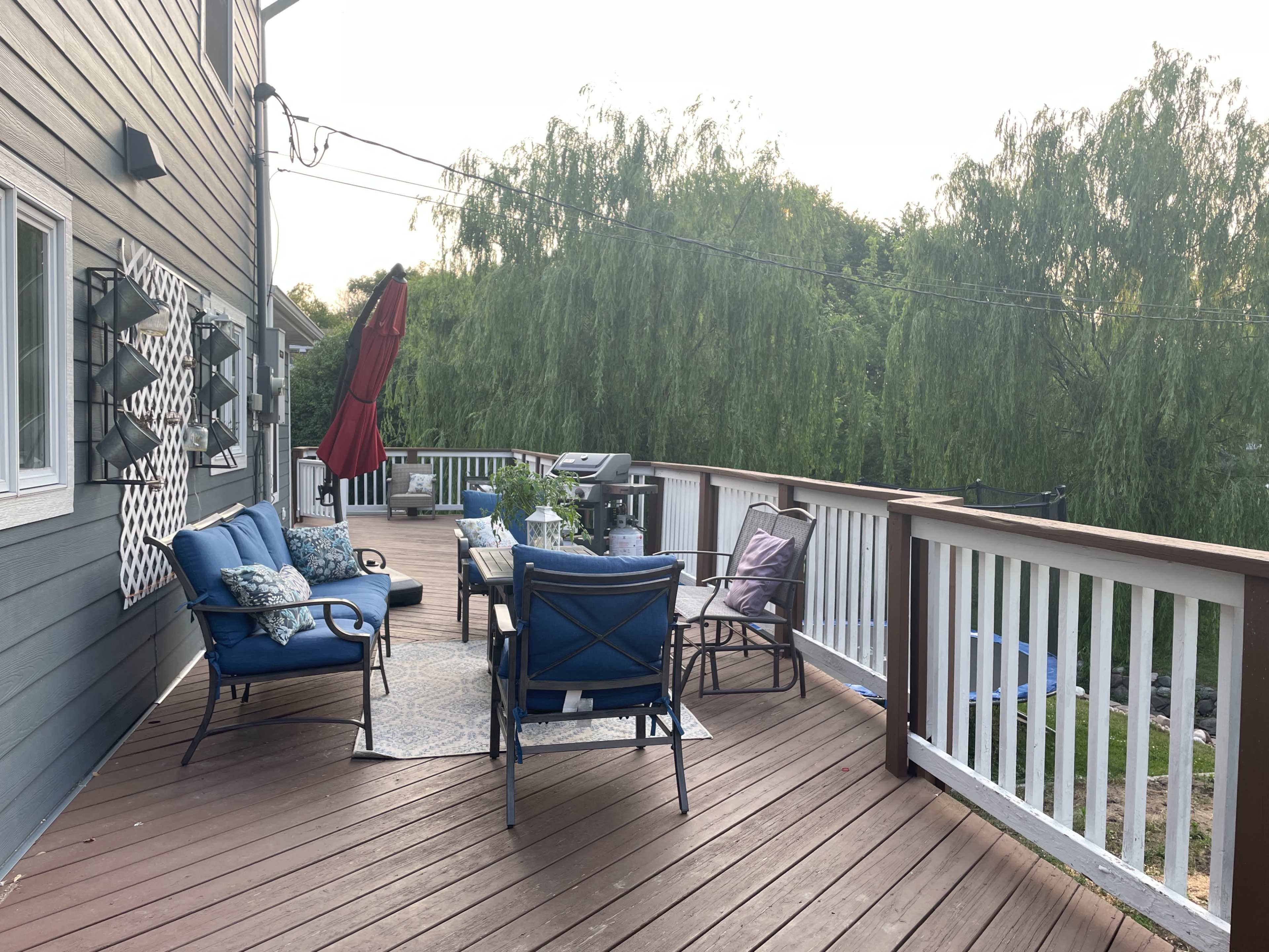 The image shows a wooden deck with blue seating, a table, and a red umbrella, surrounded by greenery.
