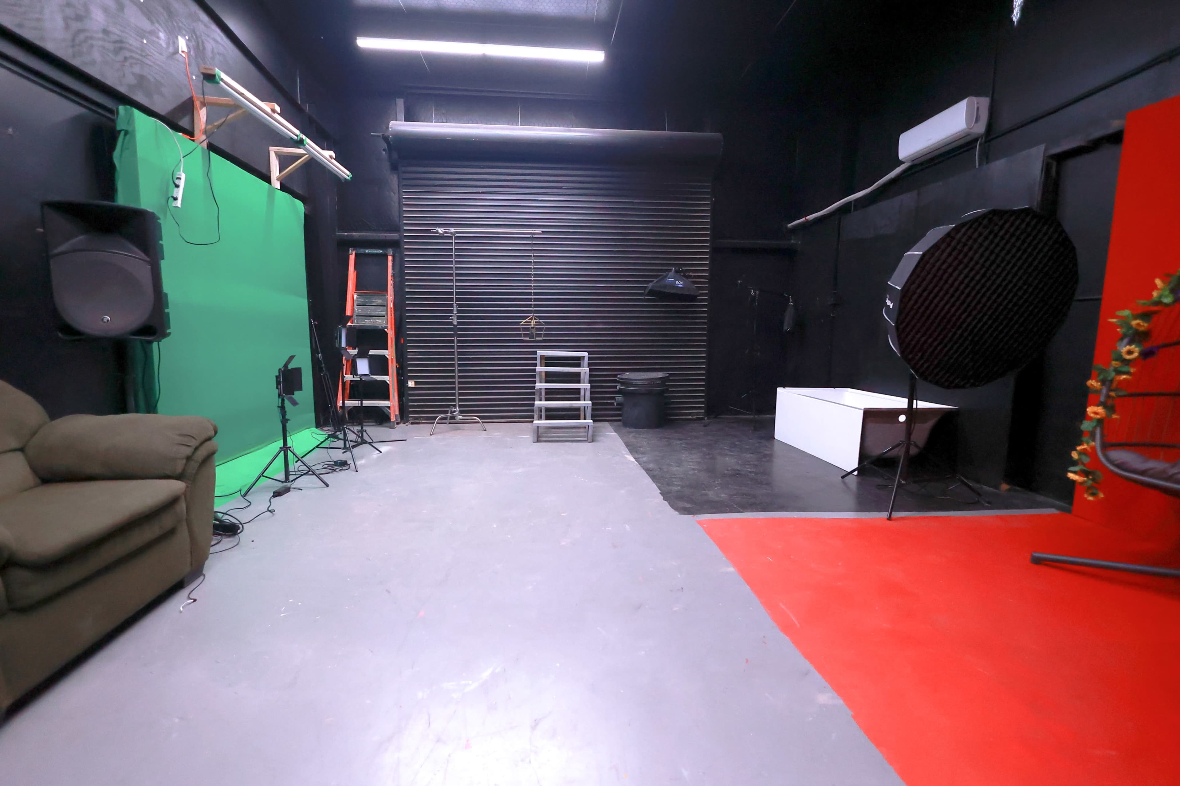 A studio space featuring a green backdrop, a red area, various lighting equipment, a ladder, and a couple of pieces of furniture.