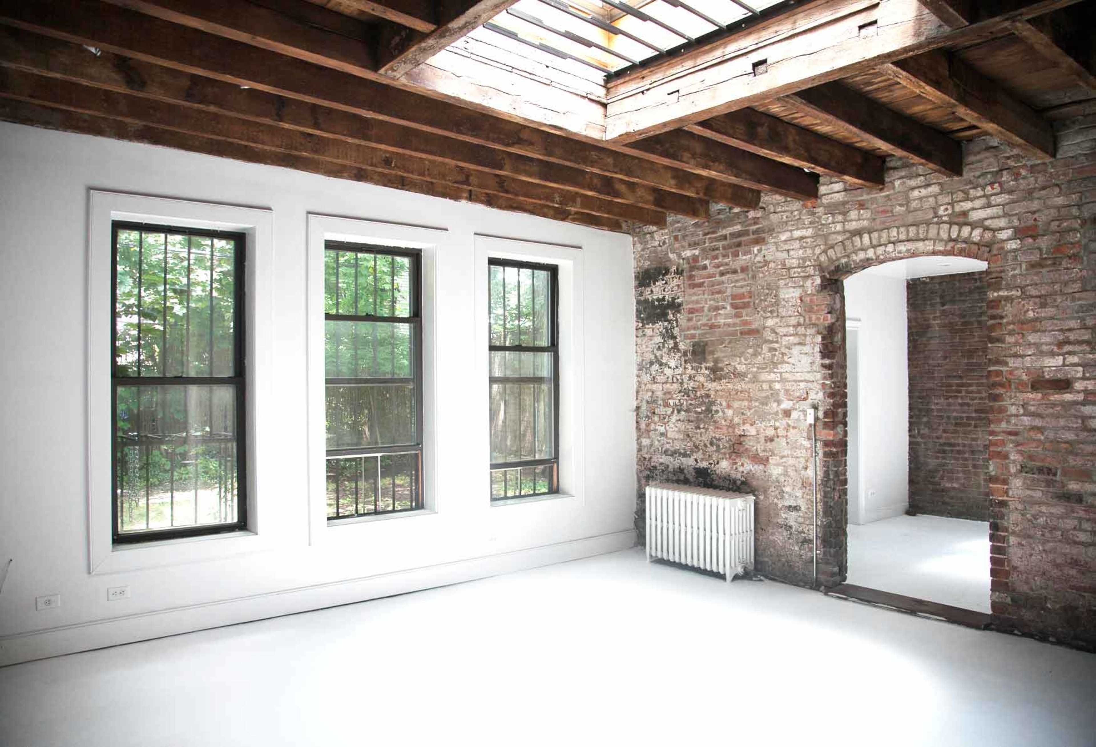 An empty room with large windows, exposed brick walls, wooden beams, and a skylight.