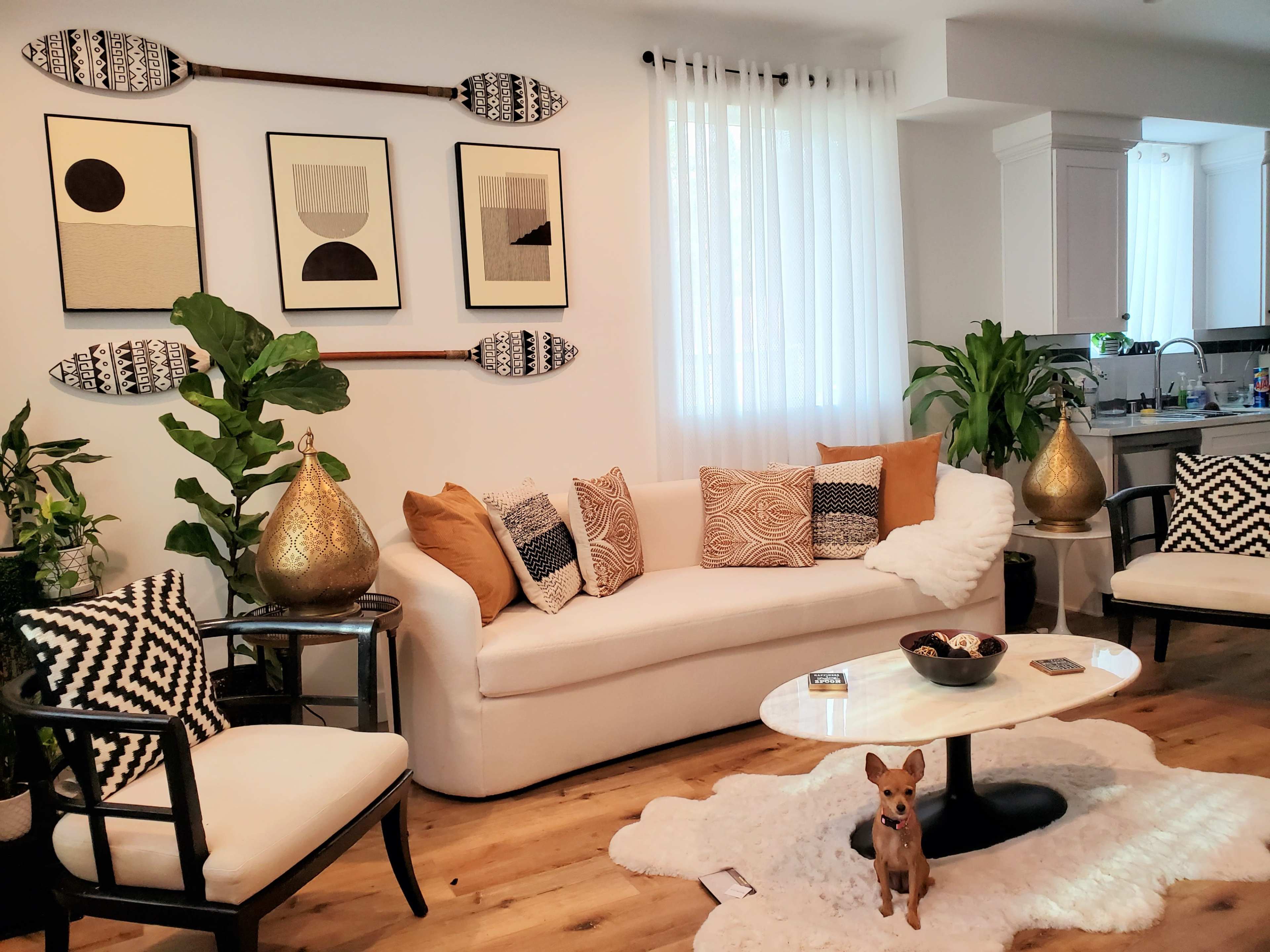 The living room features a white sofa adorned with patterned pillows, surrounded by plants and decorative artifacts under a bright, natural light from the windows.