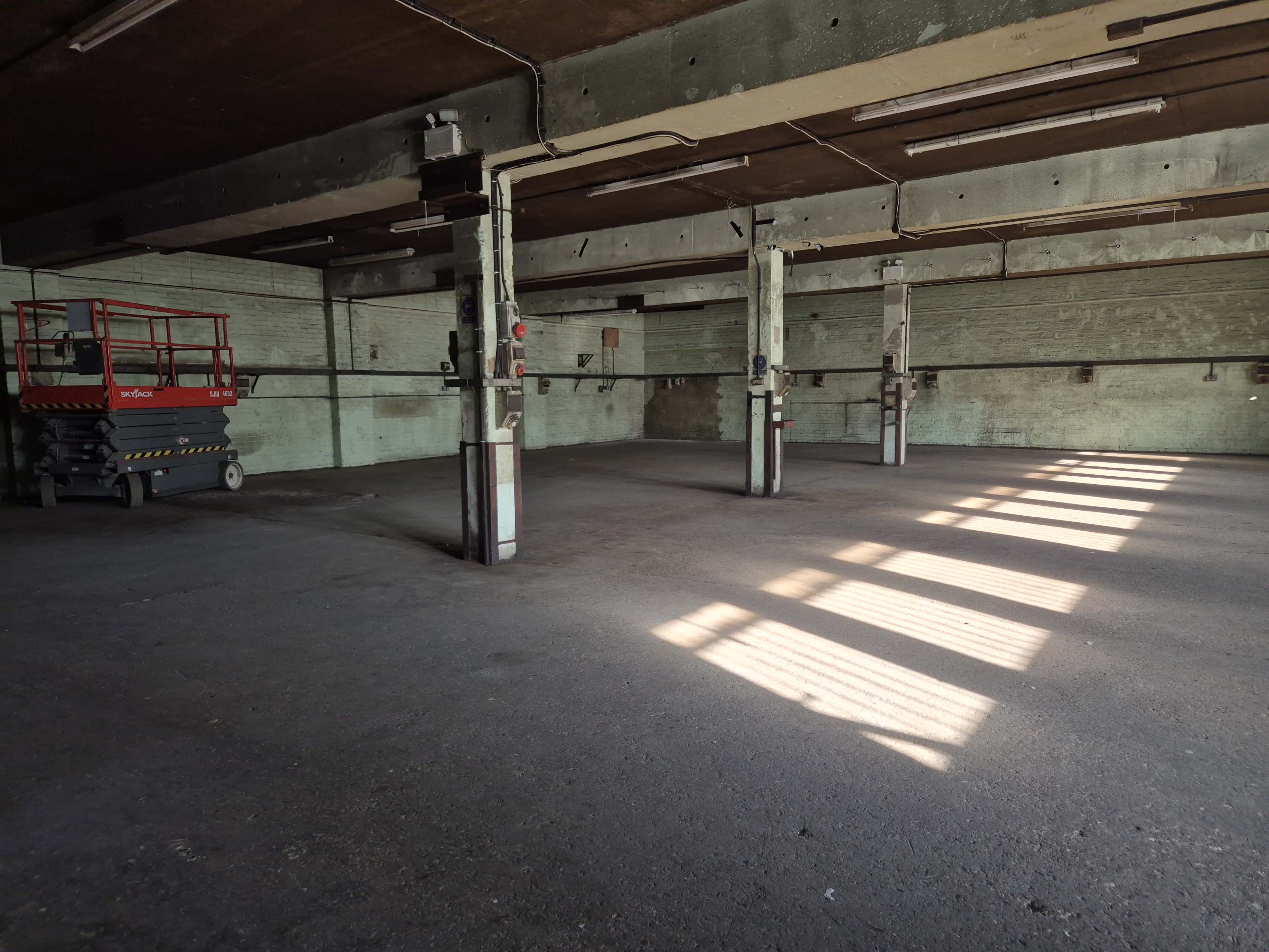 The image shows an empty warehouse with concrete floors, tall ceilings, and sunlight casting shadows through the windows.