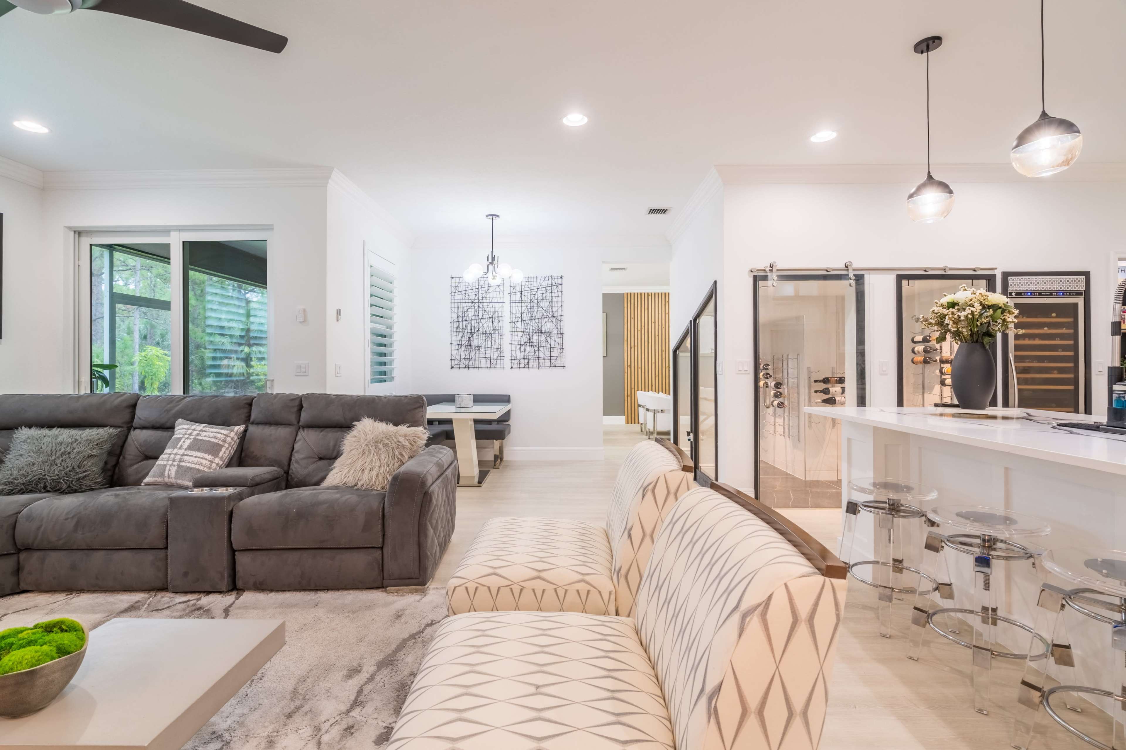 A modern living space featuring a gray sectional sofa, a patterned white couch, and a kitchen with a bar and dining area.