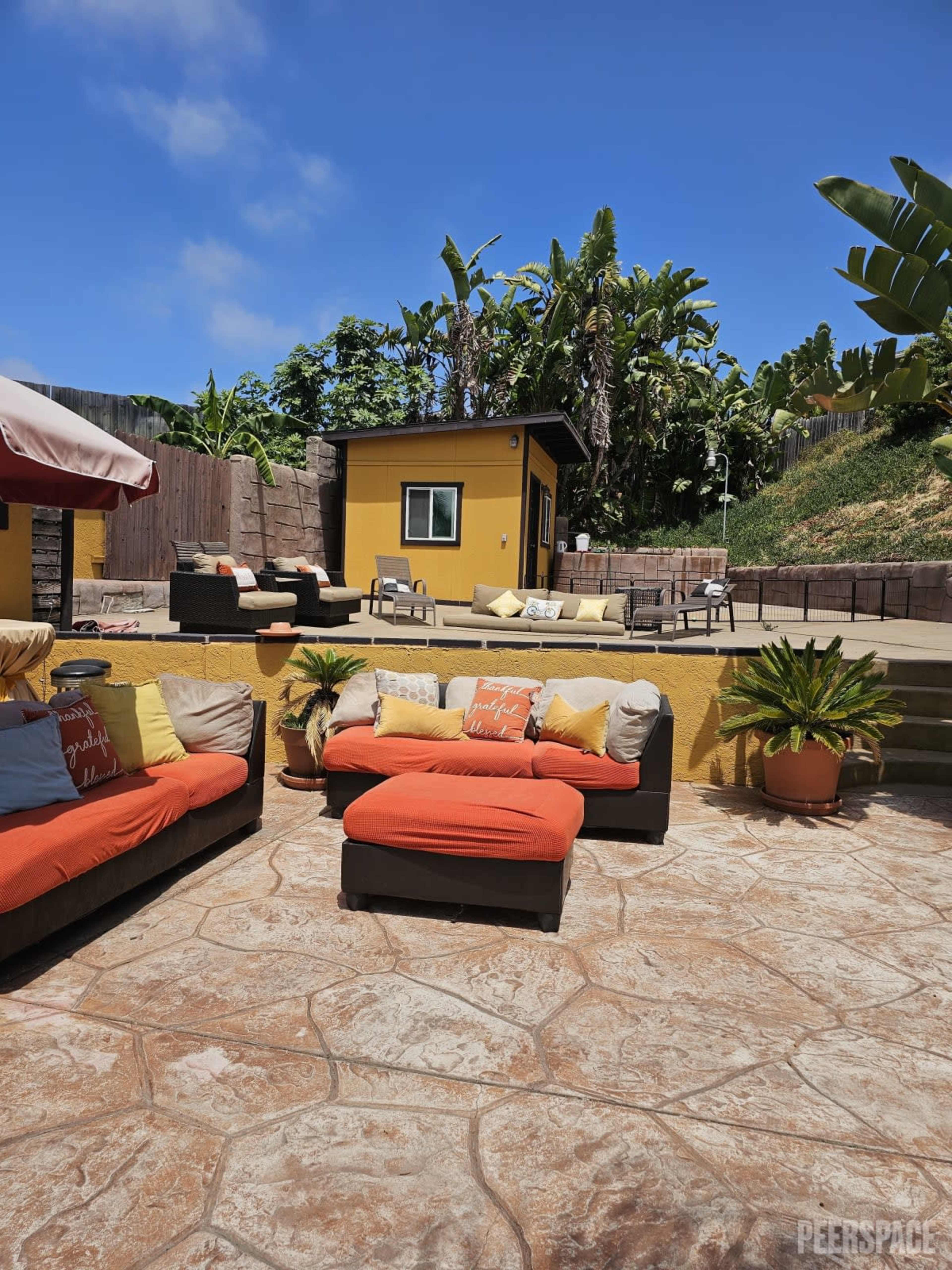 A patio area features orange and black couches surrounded by plants, with a yellow building and banana trees in the background.
