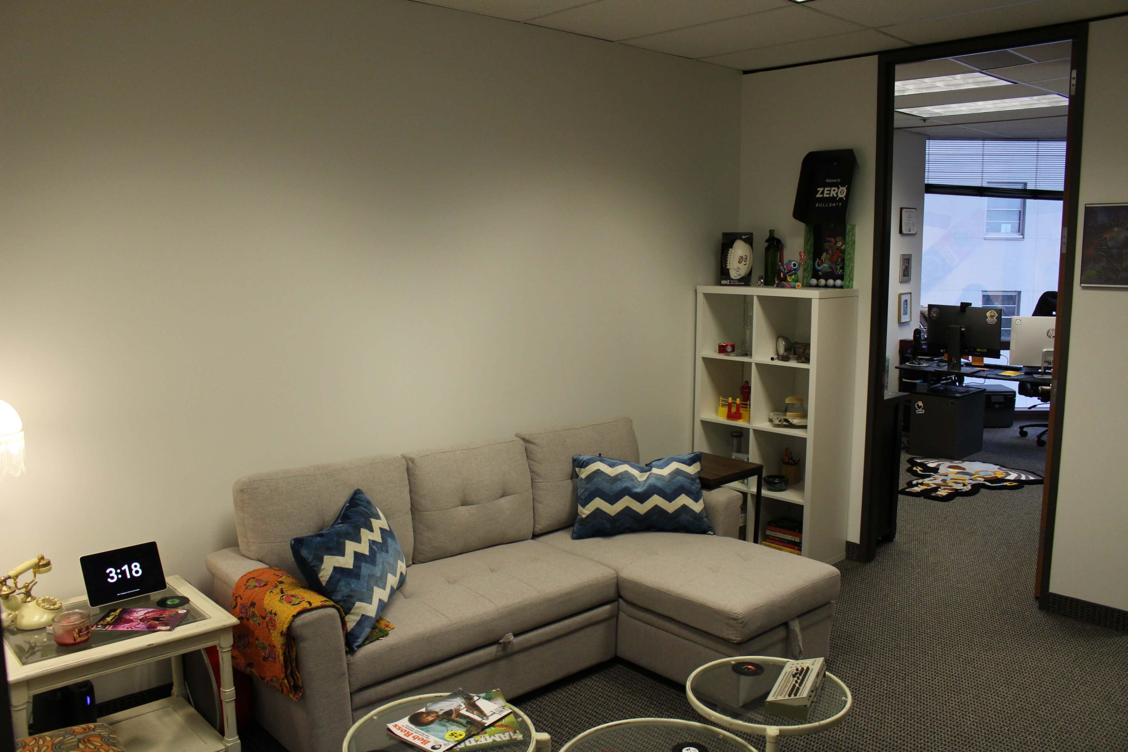 The image shows a small office space featuring a light gray sectional sofa, a clock displaying 3:18, and shelves with various decorative items and a view into an adjoining workspace.