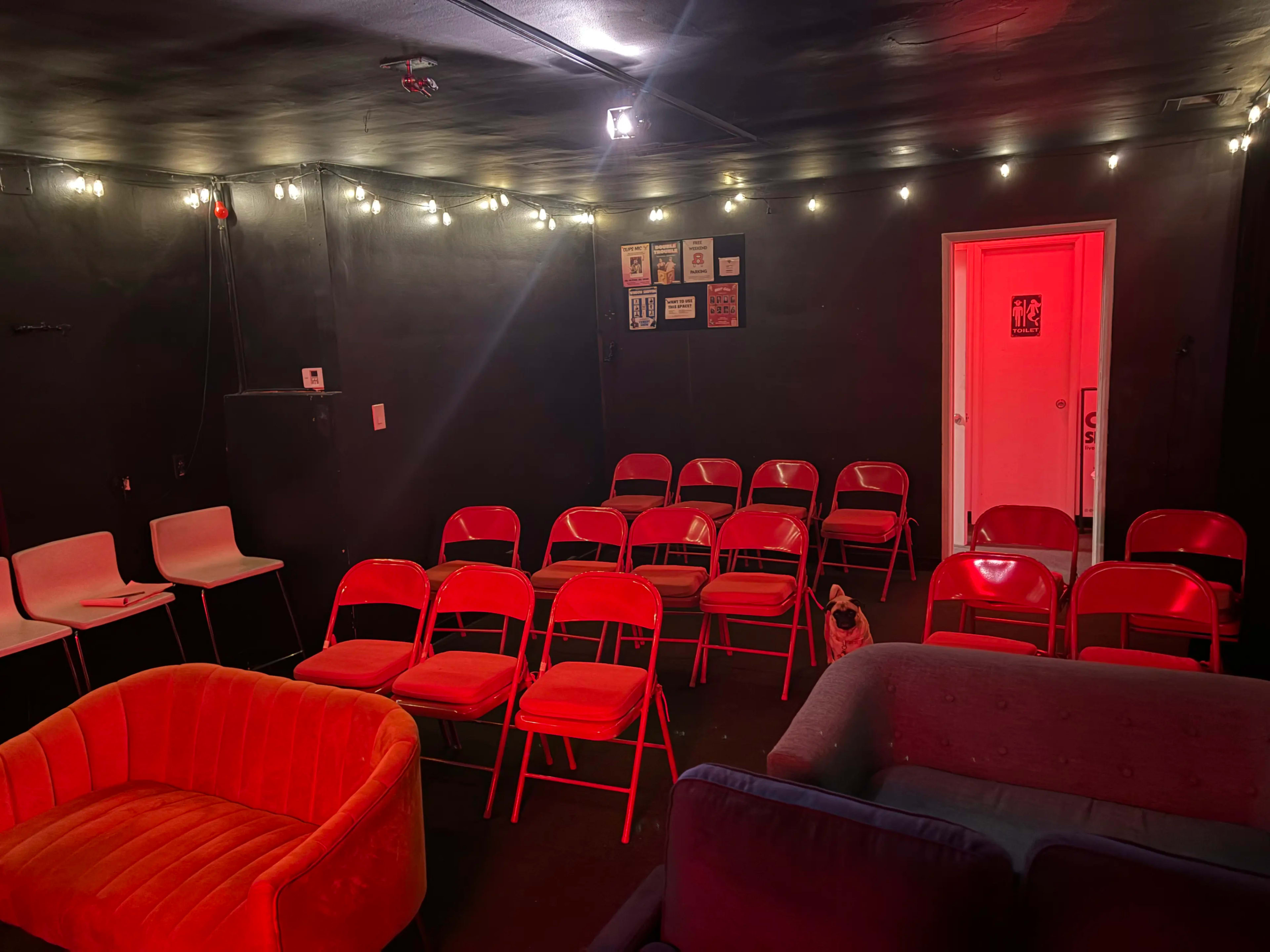 The image shows a small, dimly lit room with red chairs arranged in rows and a few couches, along with decorative string lights on the ceiling.