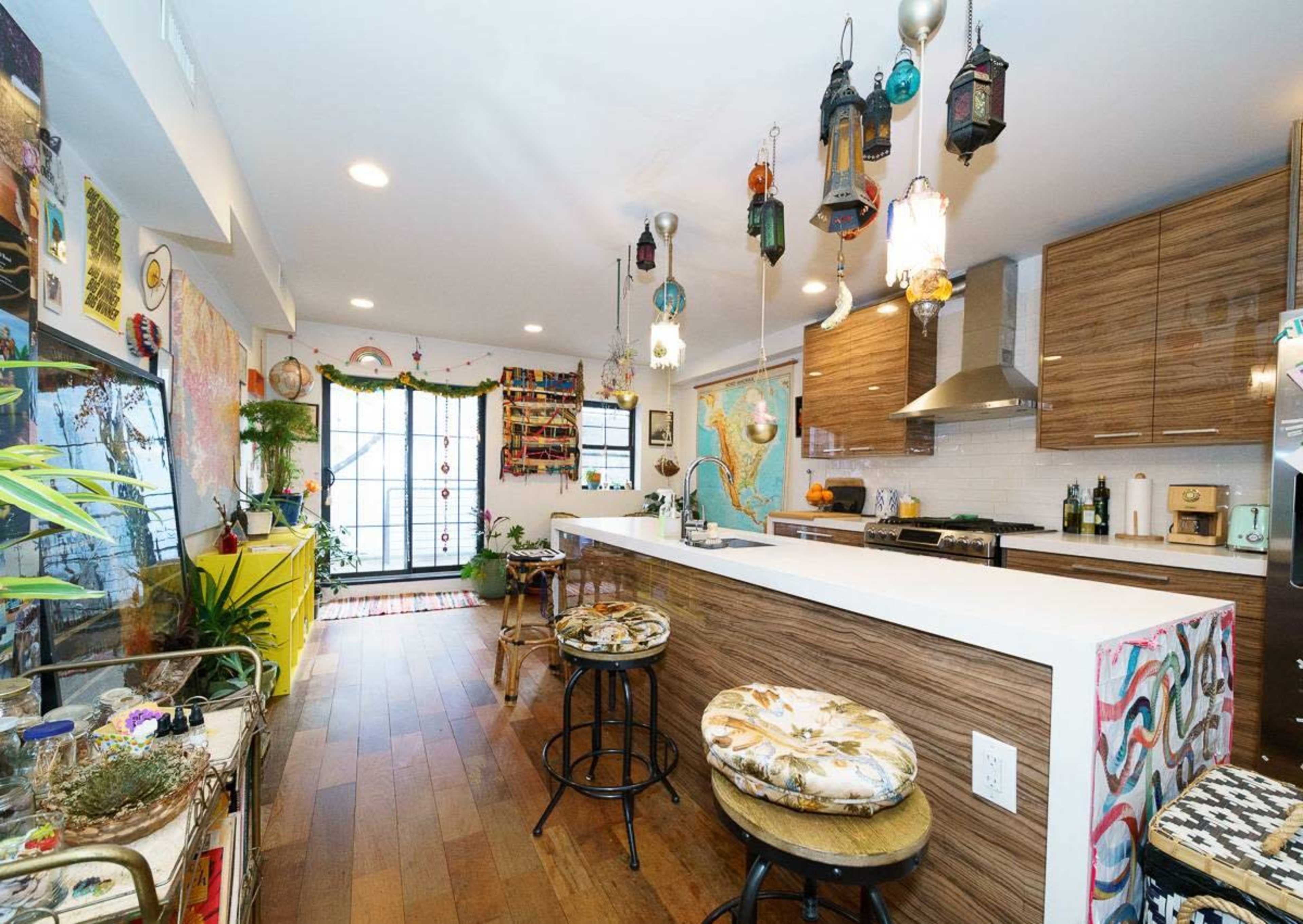 A modern kitchen with wooden cabinets, a long island countertop, and colorful decor, including hanging lanterns and various plants.