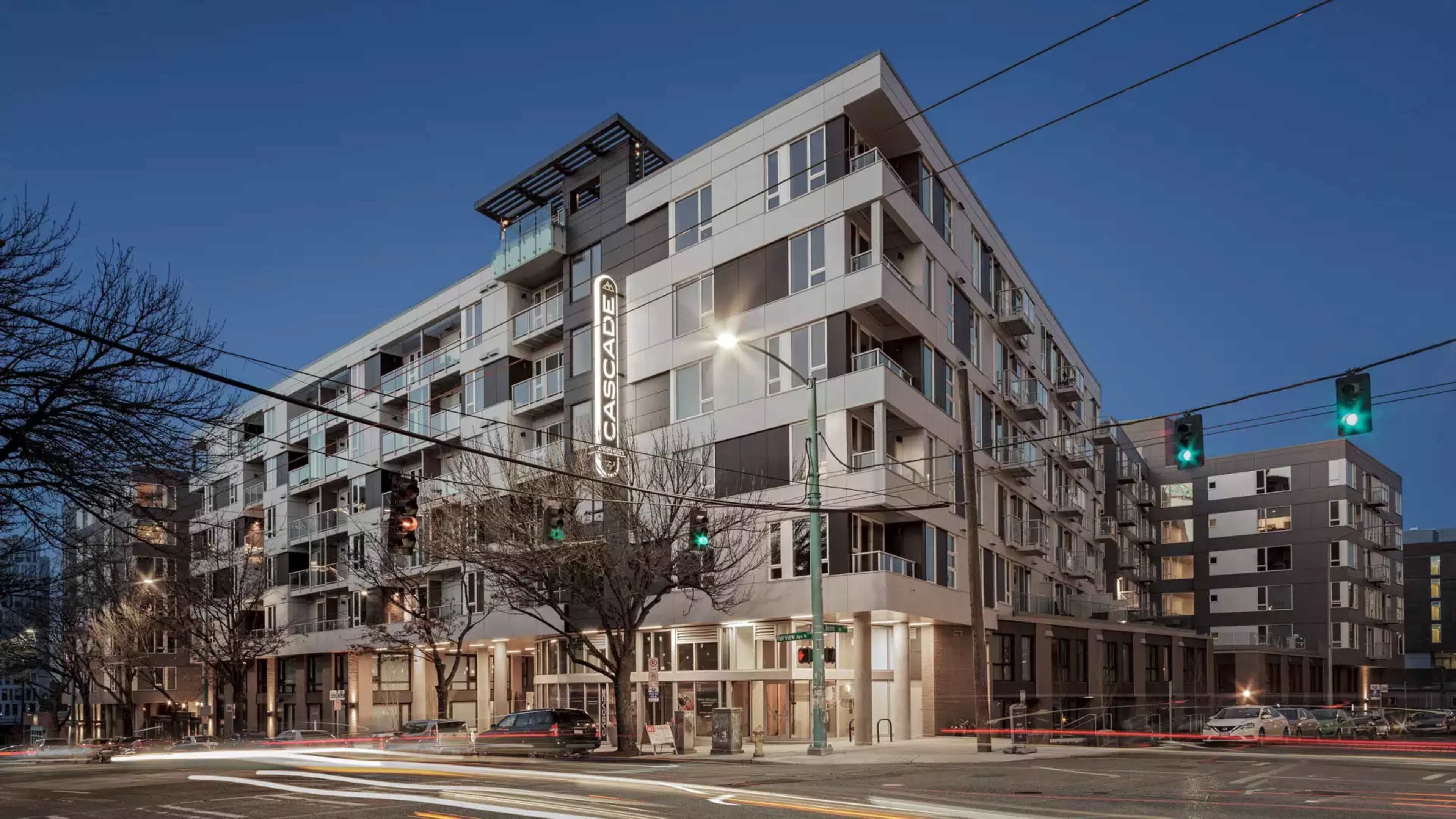 A modern residential building with large windows and a prominent sign, situated at the intersection of two streets during twilight.
