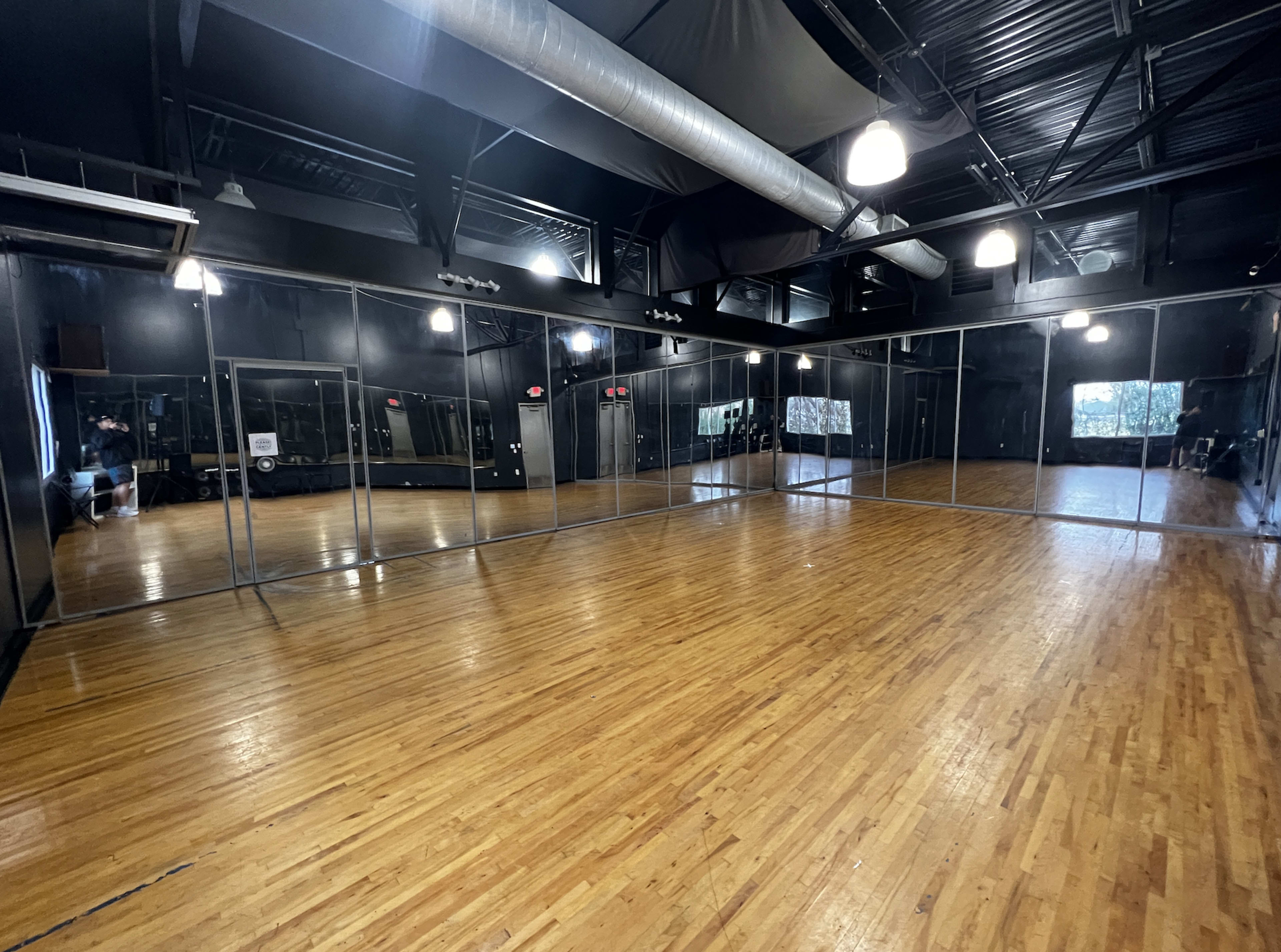 A spacious dance studio with wooden floors, large mirrors on one wall, and minimal lighting.