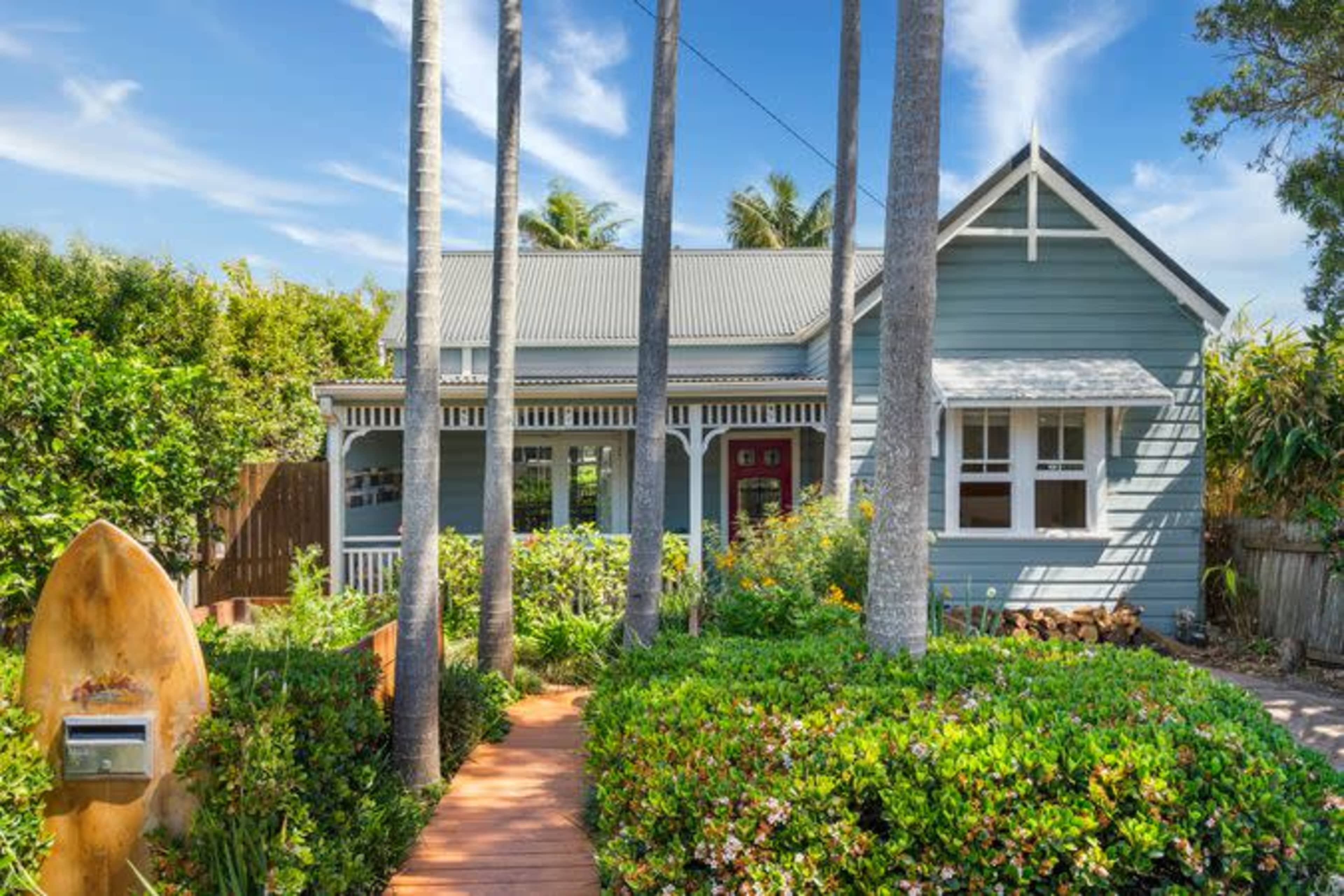 A blue wooden house with a gray roof is surrounded by palm trees and lush greenery, featuring a wooden pathway leading to the front door.