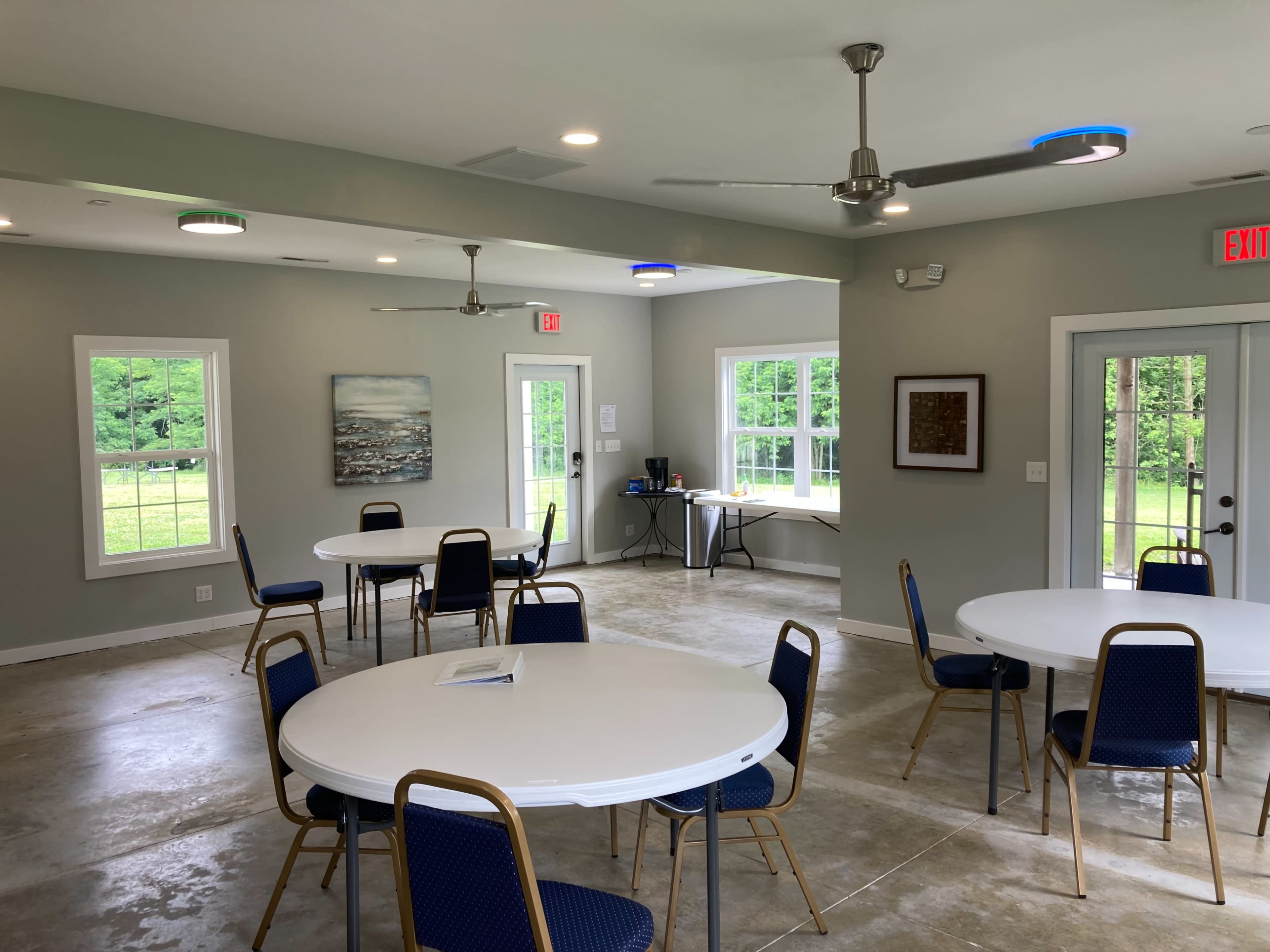An indoor meeting room with several round tables and blue chairs, featuring large windows that overlook a green landscape.