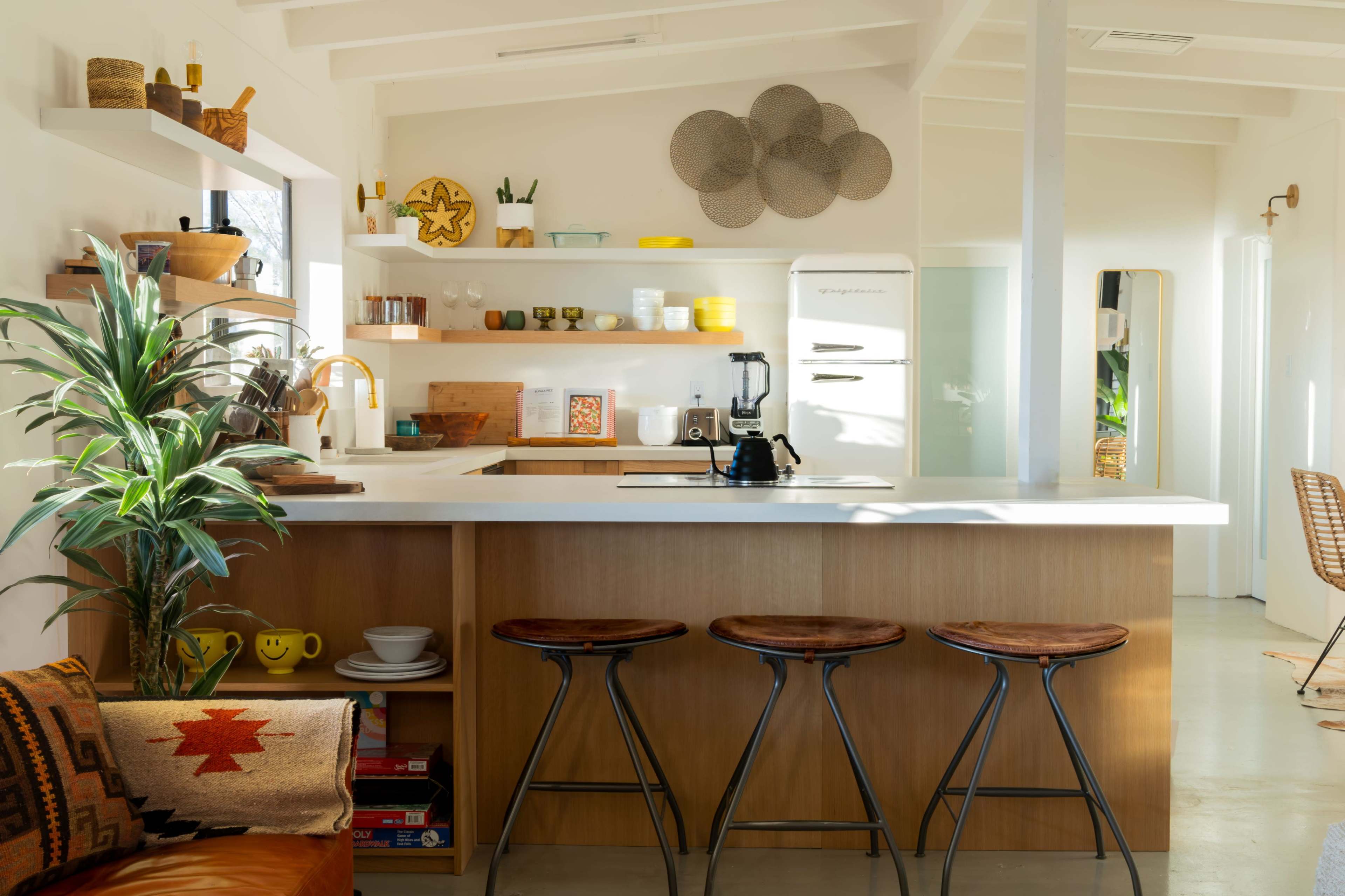 A bright and modern kitchen with a white countertop, wooden shelves filled with dishware, and three metal stools at the bar.