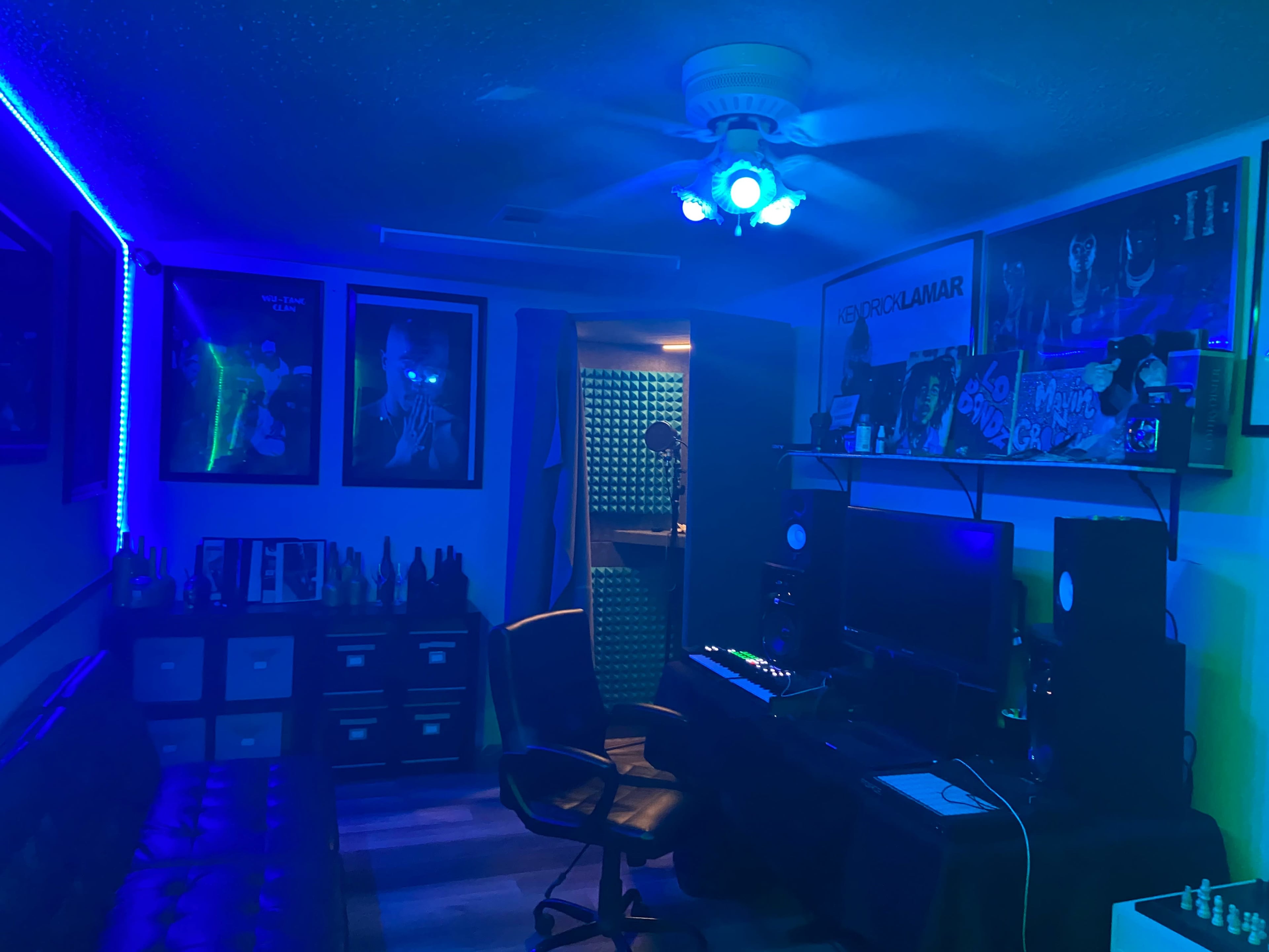 A dimly lit music studio featuring a black leather couch, a desk with audio equipment, and framed posters on the walls.
