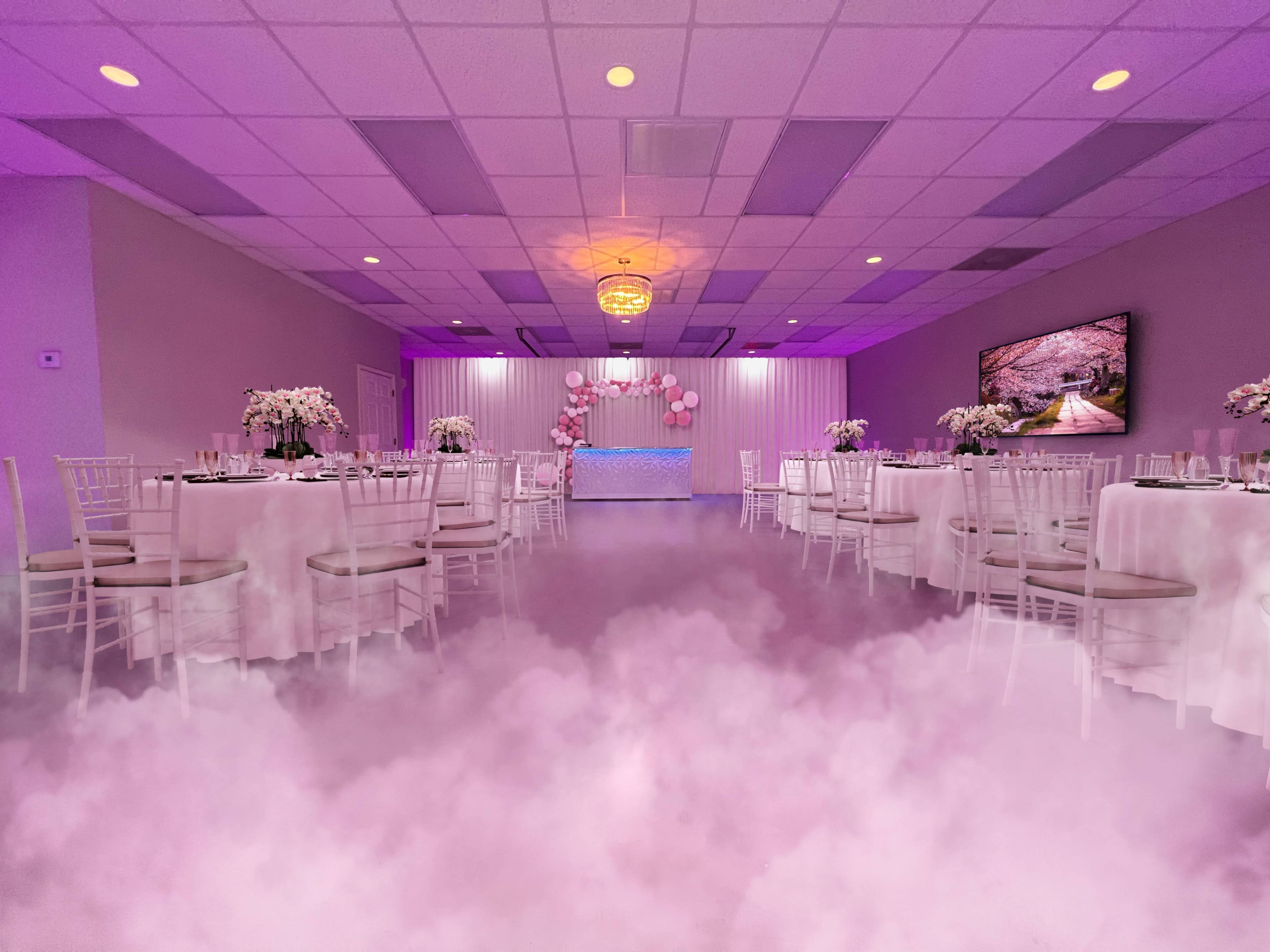 A banquet hall adorned with purple lighting features round tables draped in white, surrounded by a misty floor and a decorated backdrop at one end.