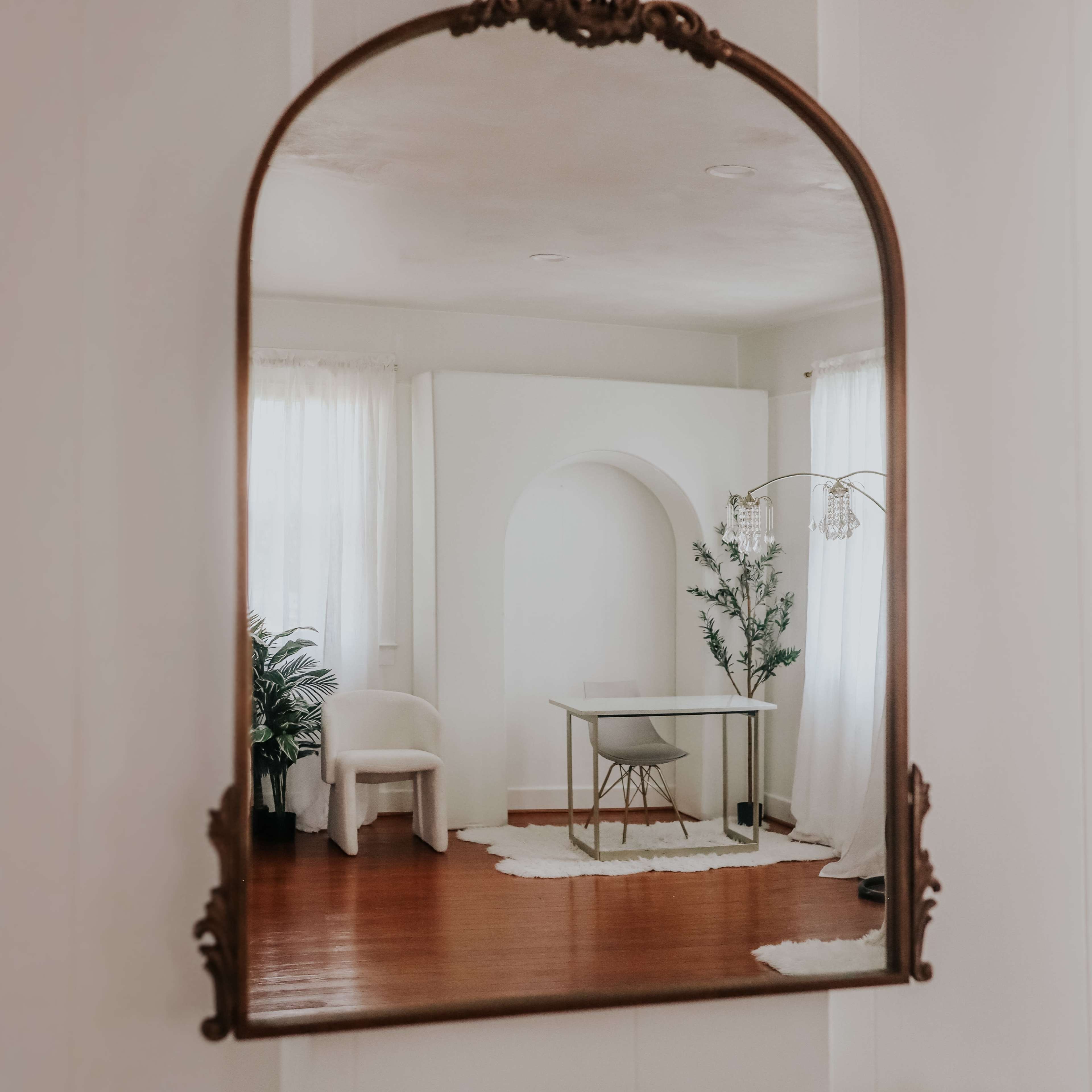 The image depicts a room with a white archway, a desk and chair, and large windows draped with white curtains, all reflected in an ornate mirror.
