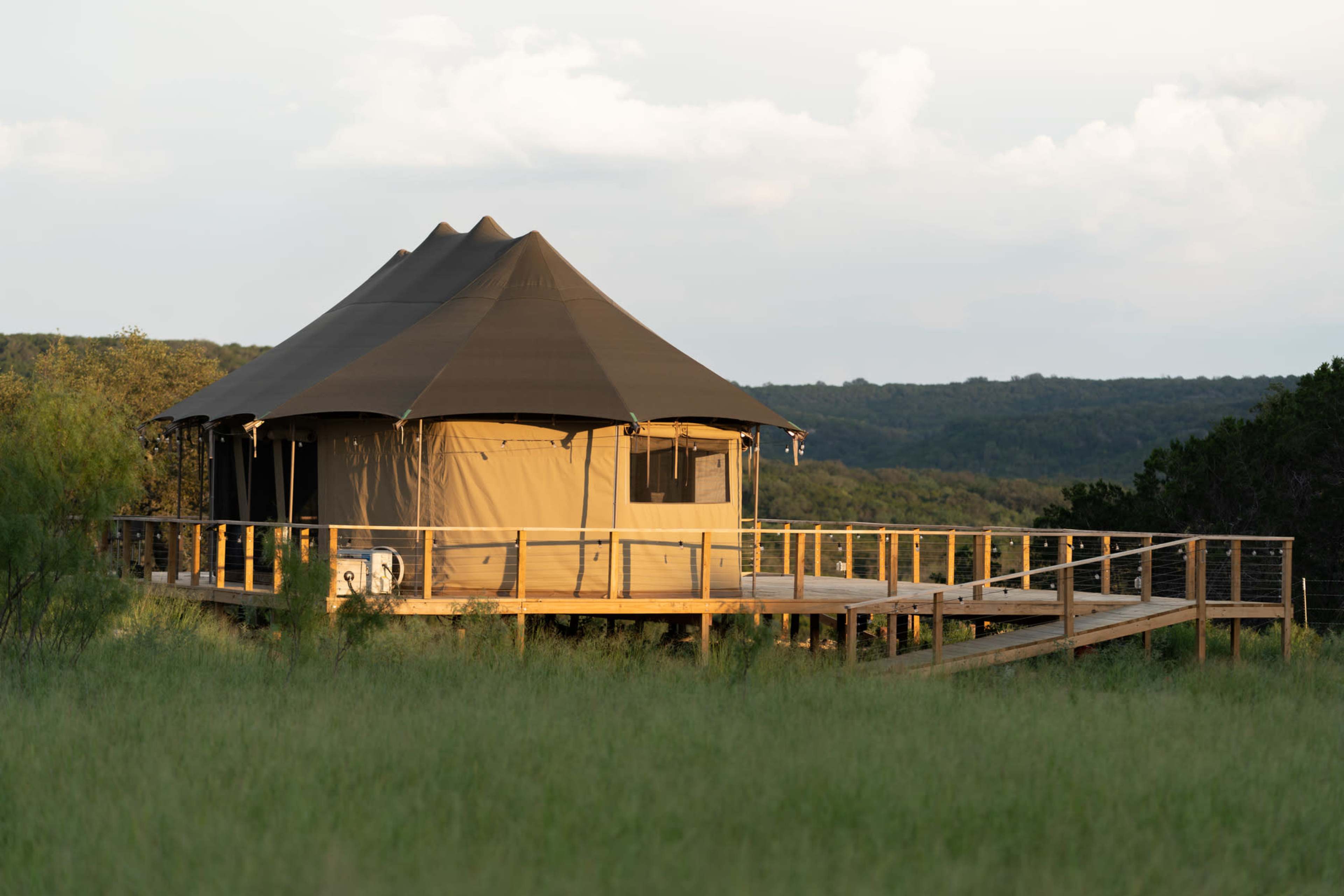 A large, tent-style structure with a conical roof is situated on a wooden deck amid green grass and trees.