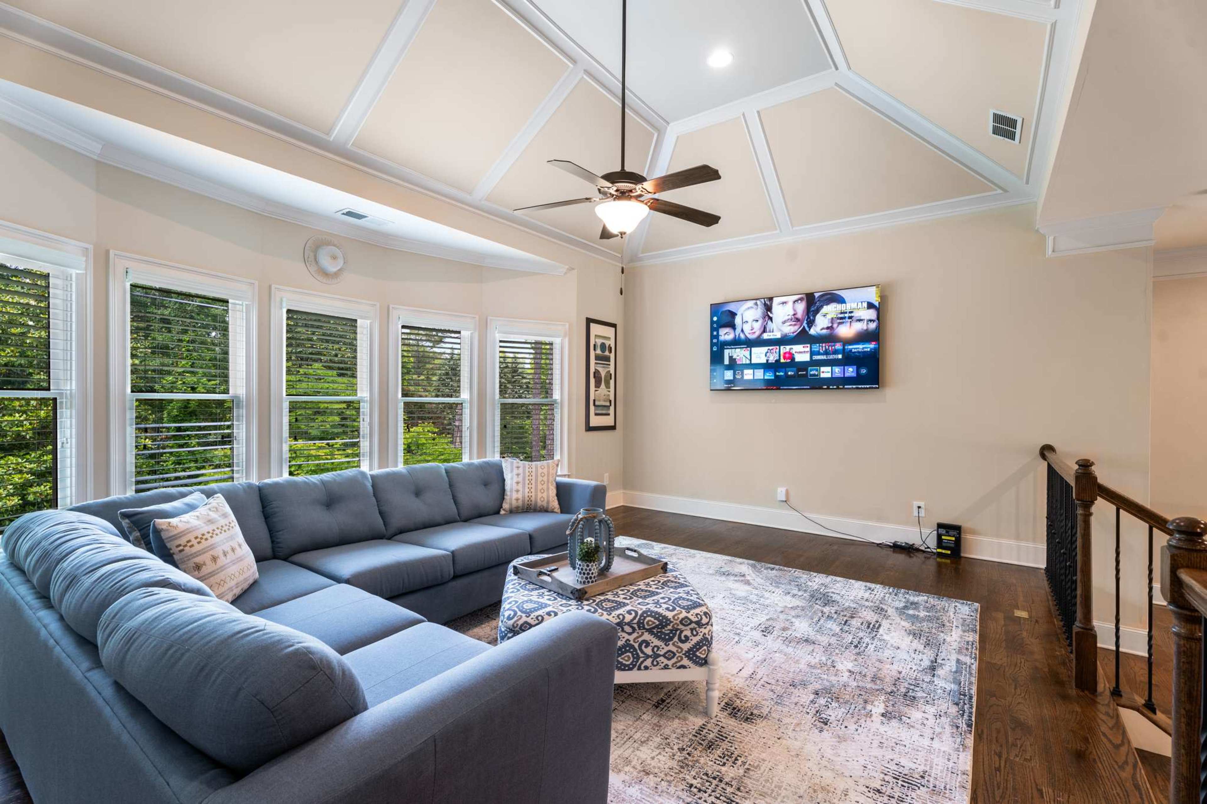 The image shows a spacious living room featuring a large sectional sofa, a coffee table, and a flat-screen TV mounted on the wall, all under a coffered ceiling with natural light streaming in through multiple windows.