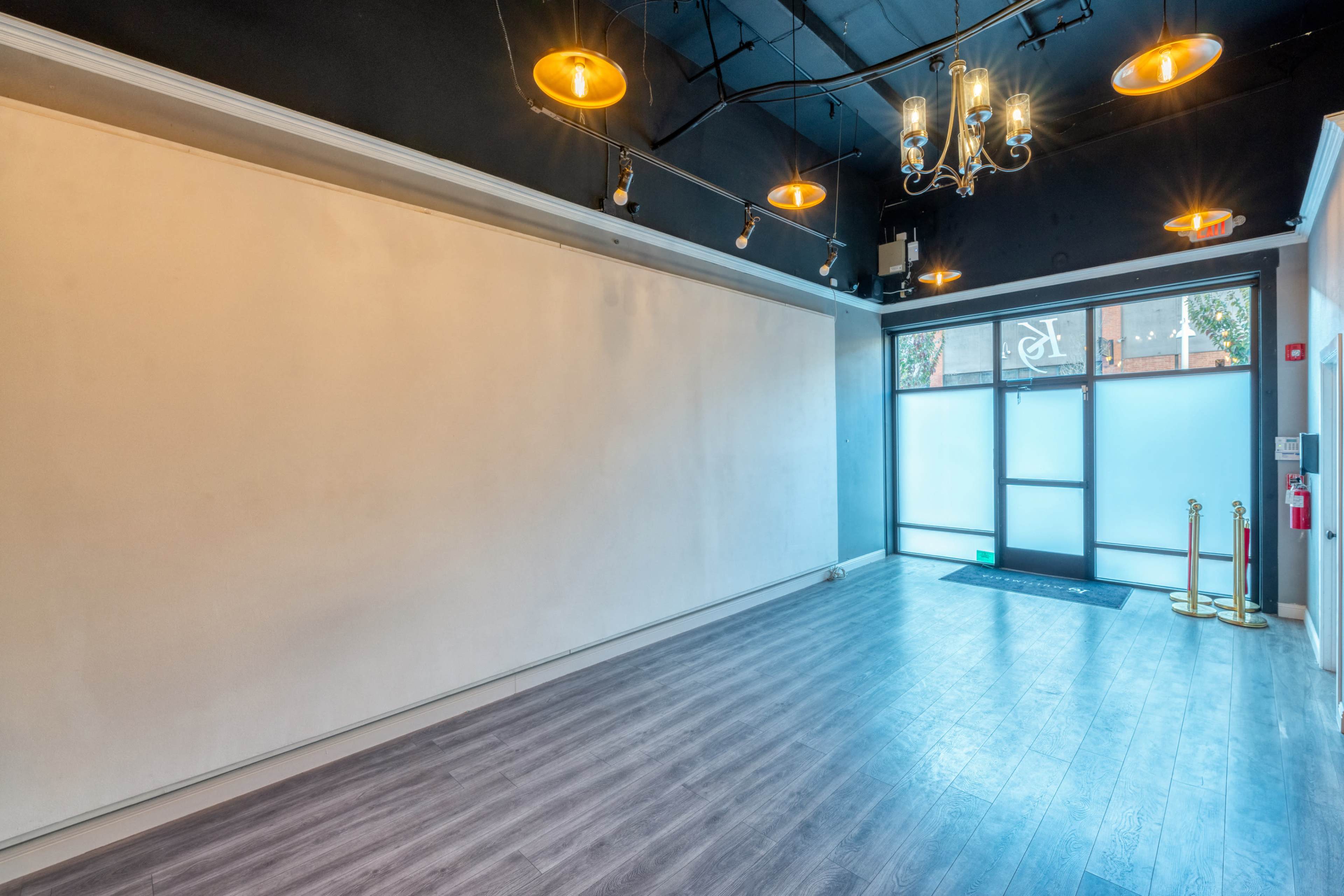 A vacant commercial space with a large, blank wall, hardwood flooring, and a glass entrance door illuminated by pendant lights.
