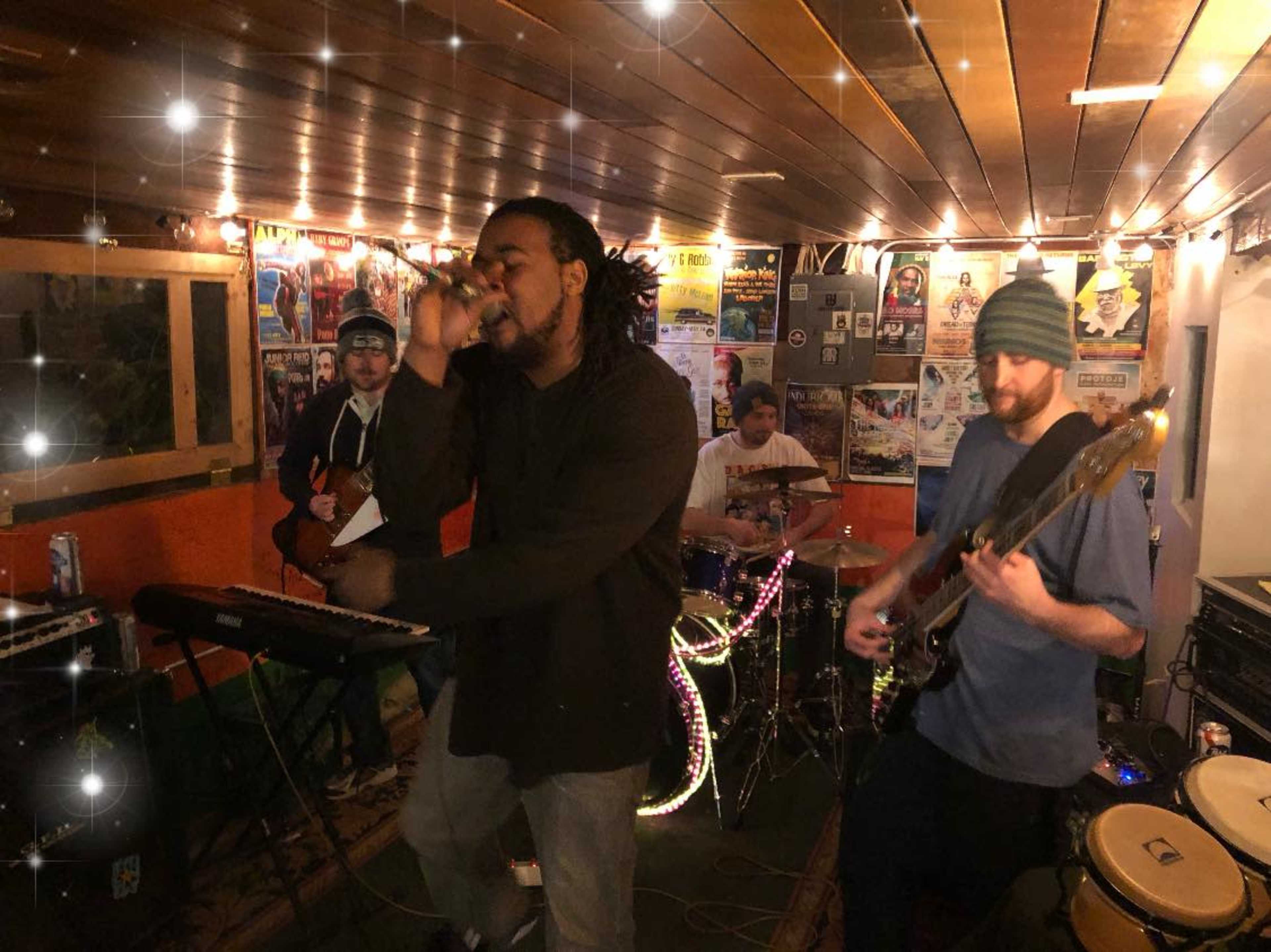 A musical group performs on stage in an intimate venue with instruments and sound equipment visible around them.